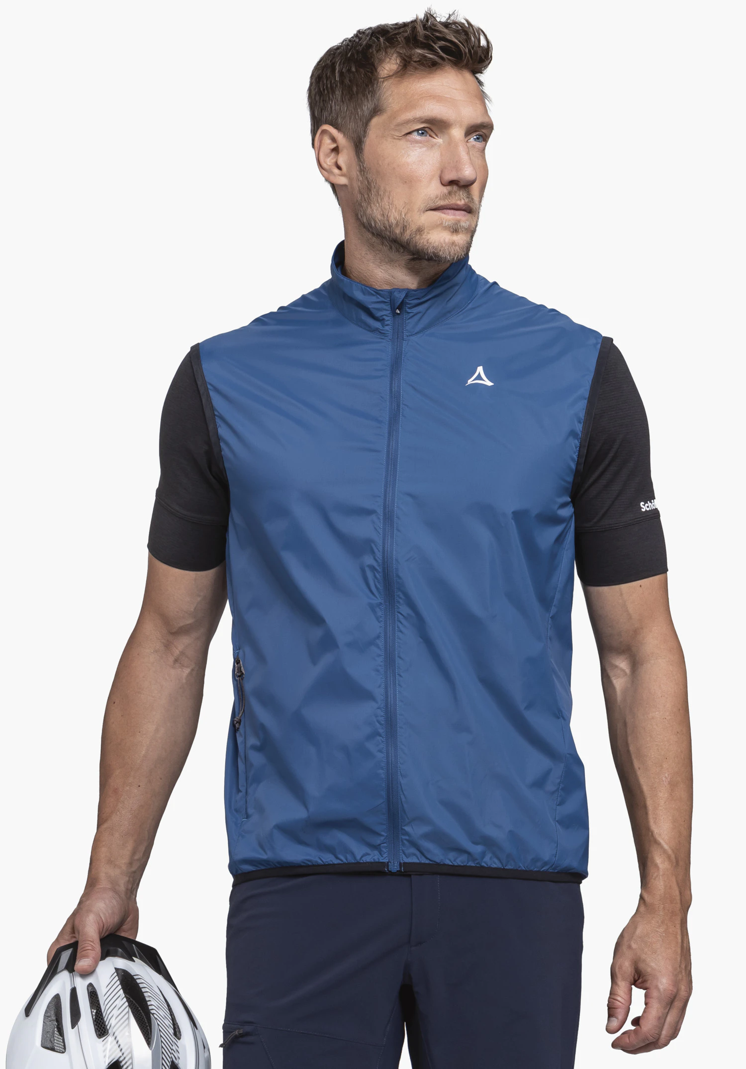 Vest Style Cannobio MNS: A model stands frontally and looks to the side. He is wearing the vest in blue with a visible Schöffel logo on the chest and holds a bicycle helmet in his hand, in front of a neutral background.