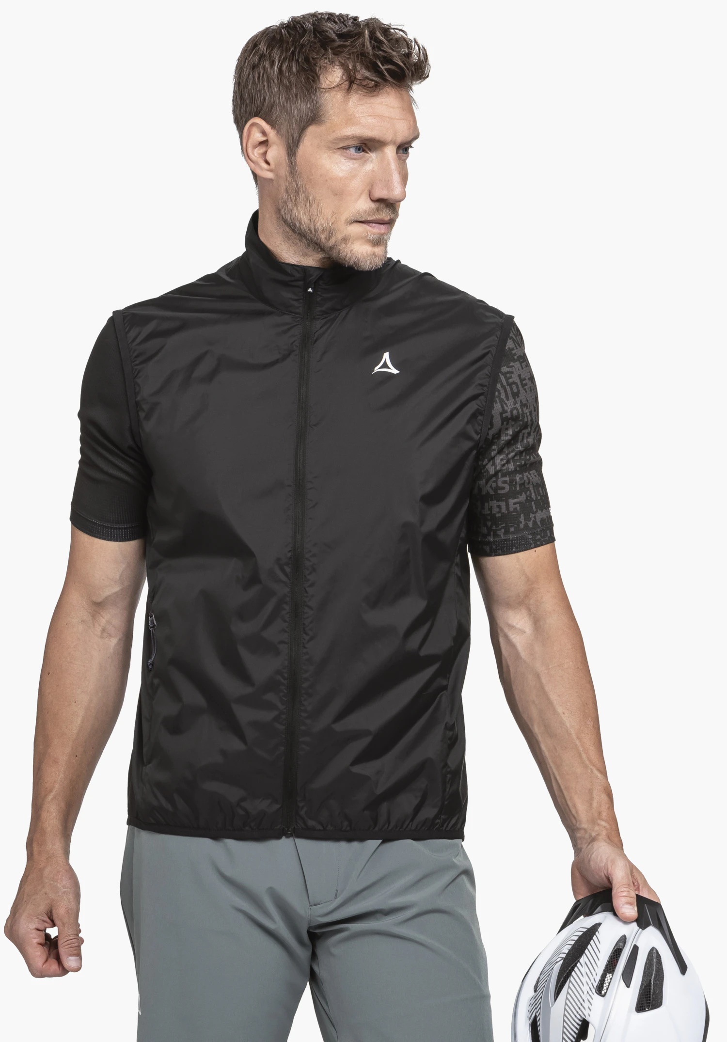 Vest Style Cannobio MNS on a male model standing slightly to the side and looking away. He is wearing the black gilet with a visible Schöffel logo on the left chest and holds a cycle helmet in front of a neutral background.