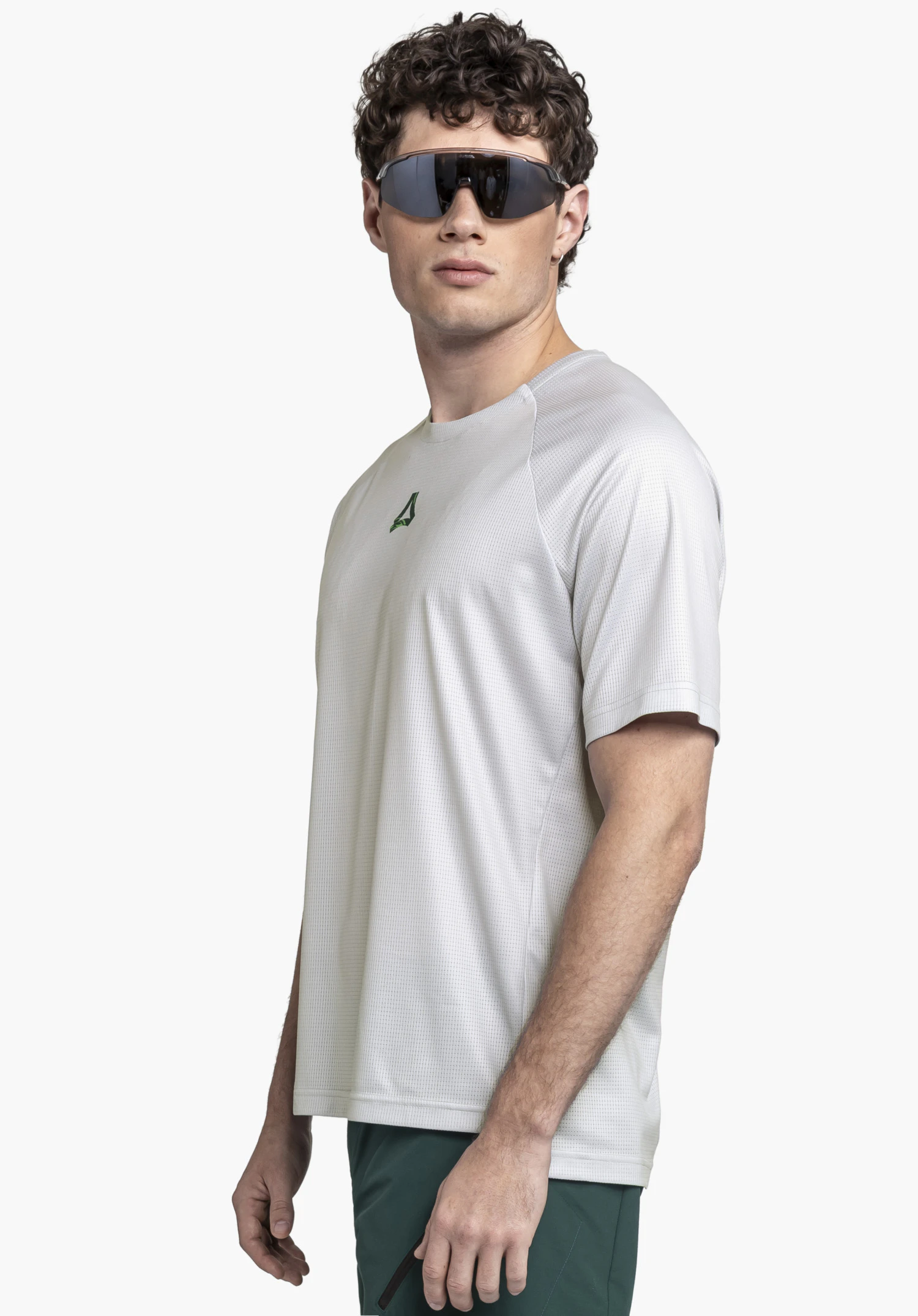 Shirt Style Koitere MEN en gris. Le modèle pose de profil devant un fond neutre et porte le t-shirt avec le logo Schöffel vert distinctif sur la poitrine. Le look sportif est complété par des lunettes de soleil.