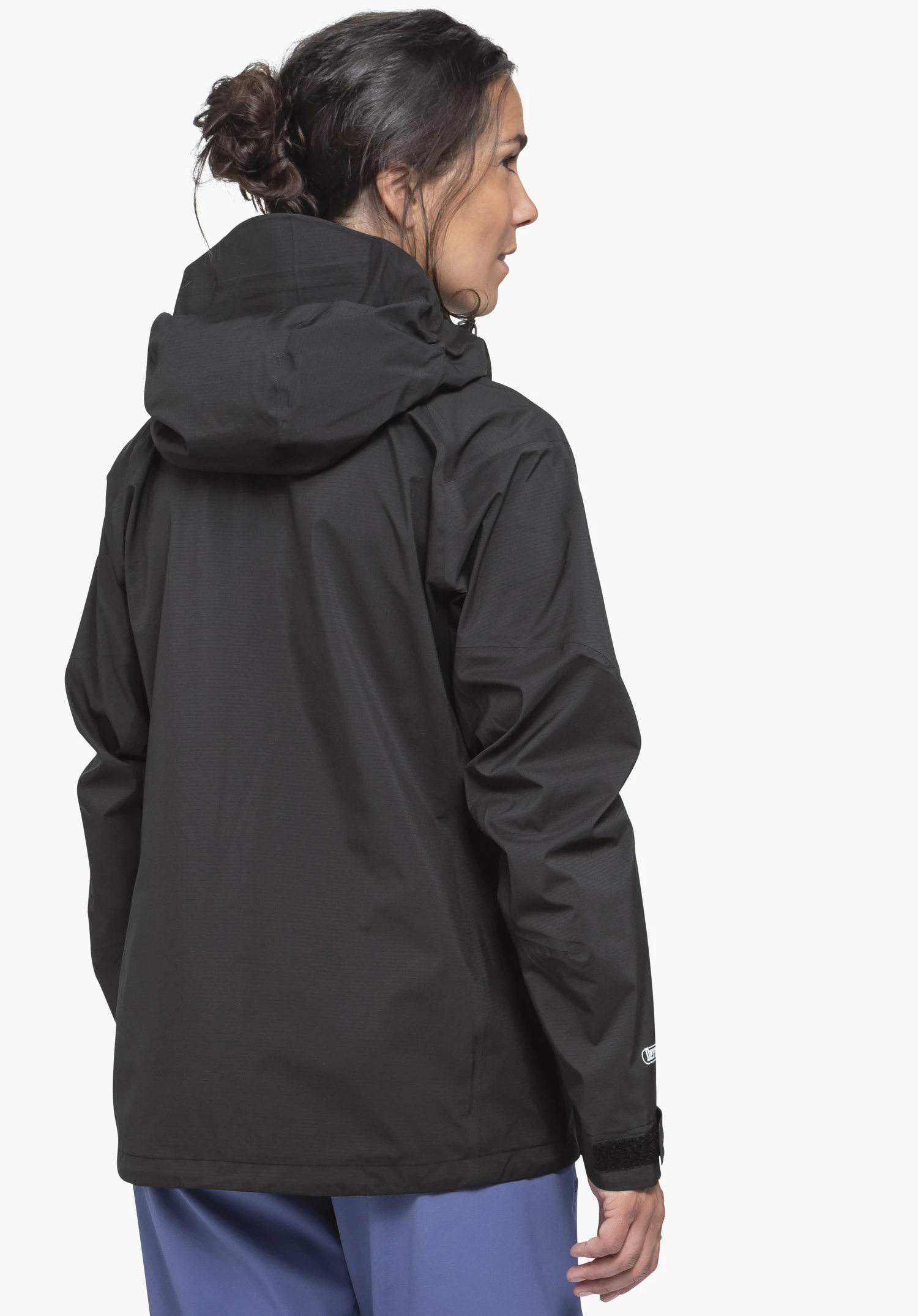 Jacket Style Blaueis WMS black rain jacket worn by a woman; she stands with back to the camera, hood visible; white background, blue trousers visible.