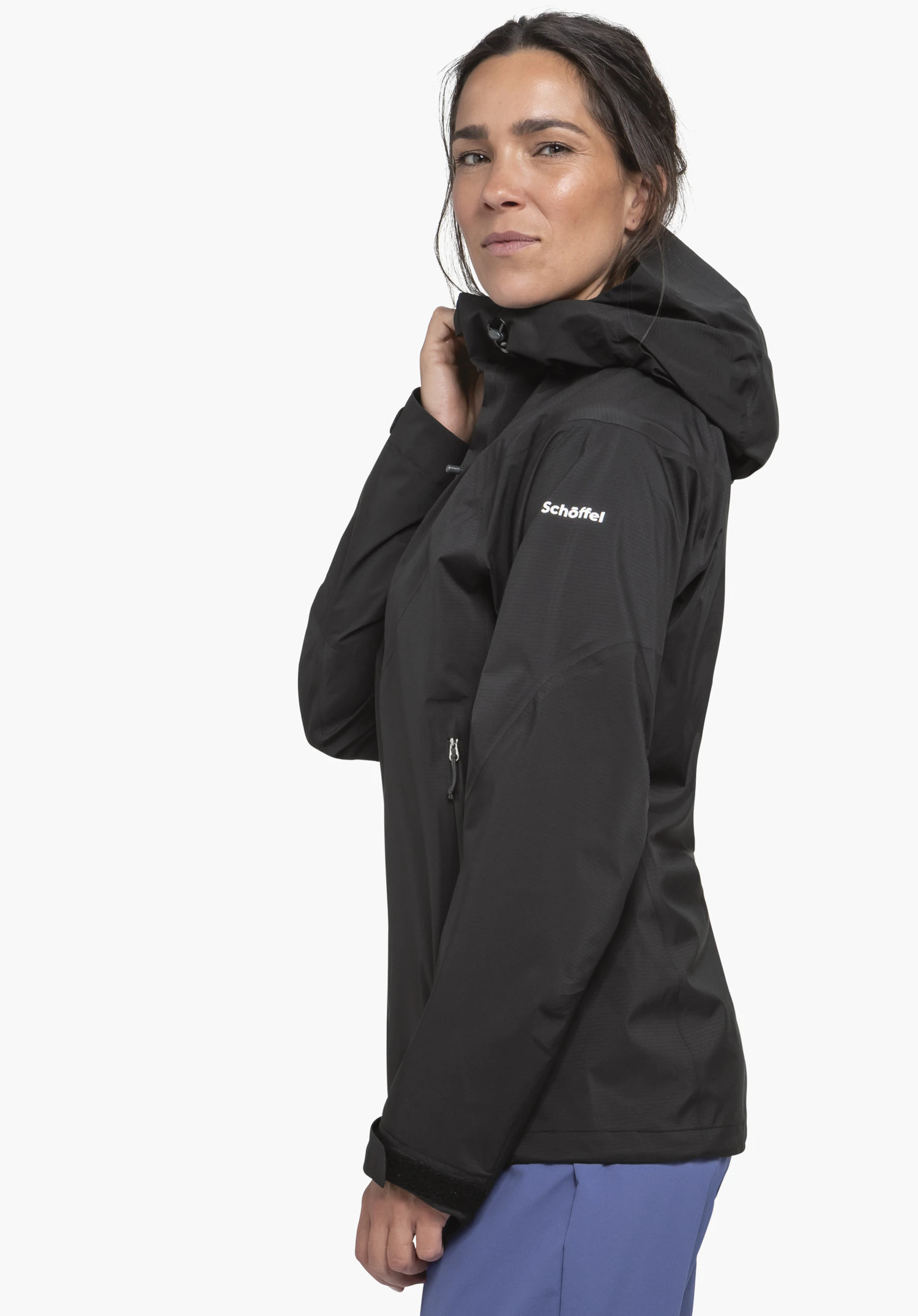 Jacket Style Blaueis WMS black outdoor jacket worn by a woman holding the hood, standing side-on in front of a white background; sleeve text: "Schöffel". Light blue trousers visible.