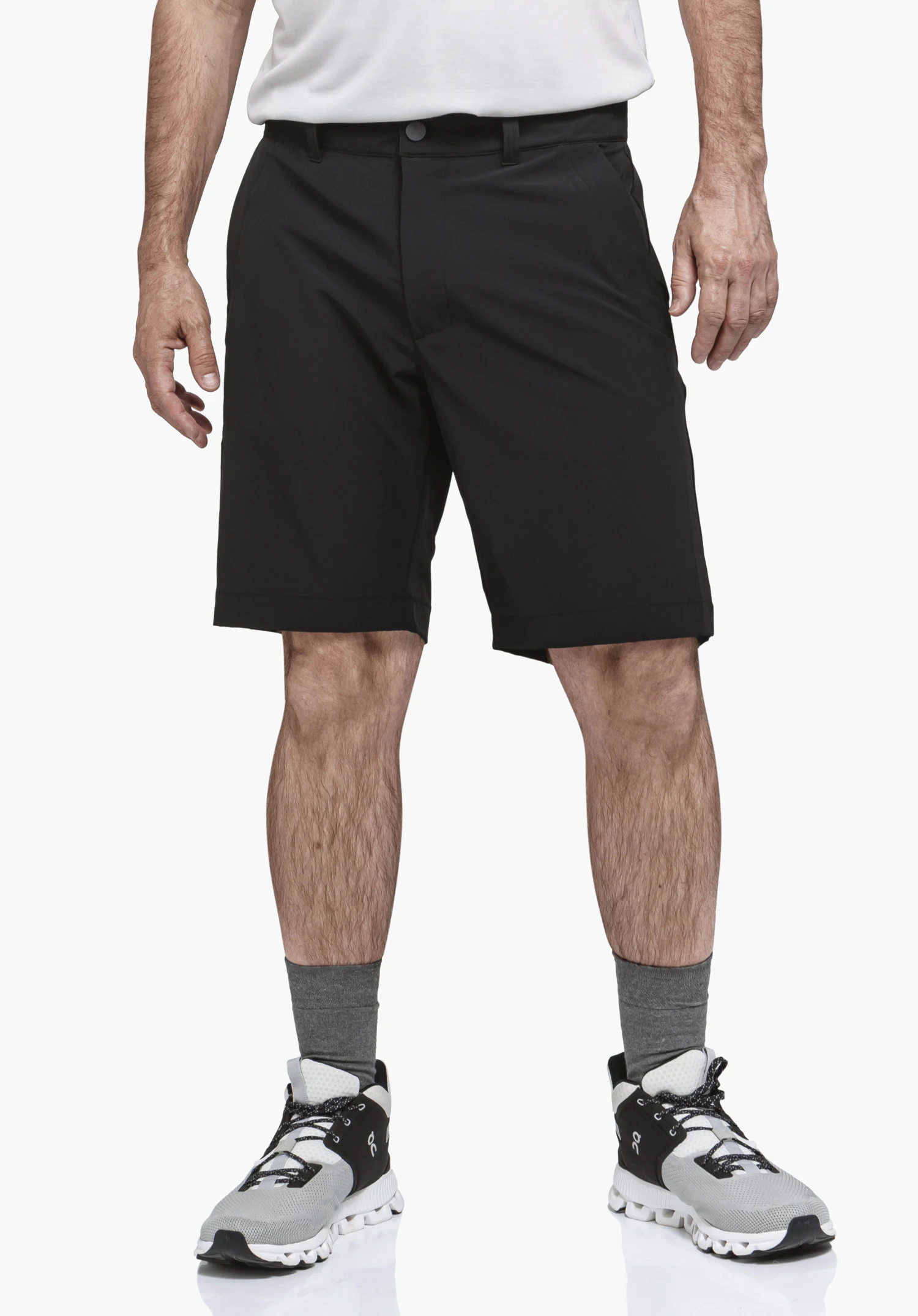 Shorts Style Gharra MNS. Ein Model steht frontal und präsentiert die schwarze Hose in der Vorderansicht. Das Outfit wird durch ein helles Oberteil, graue Socken und Sportschuhe ergänzt, fotografiert vor einem neutralen Hintergrund.