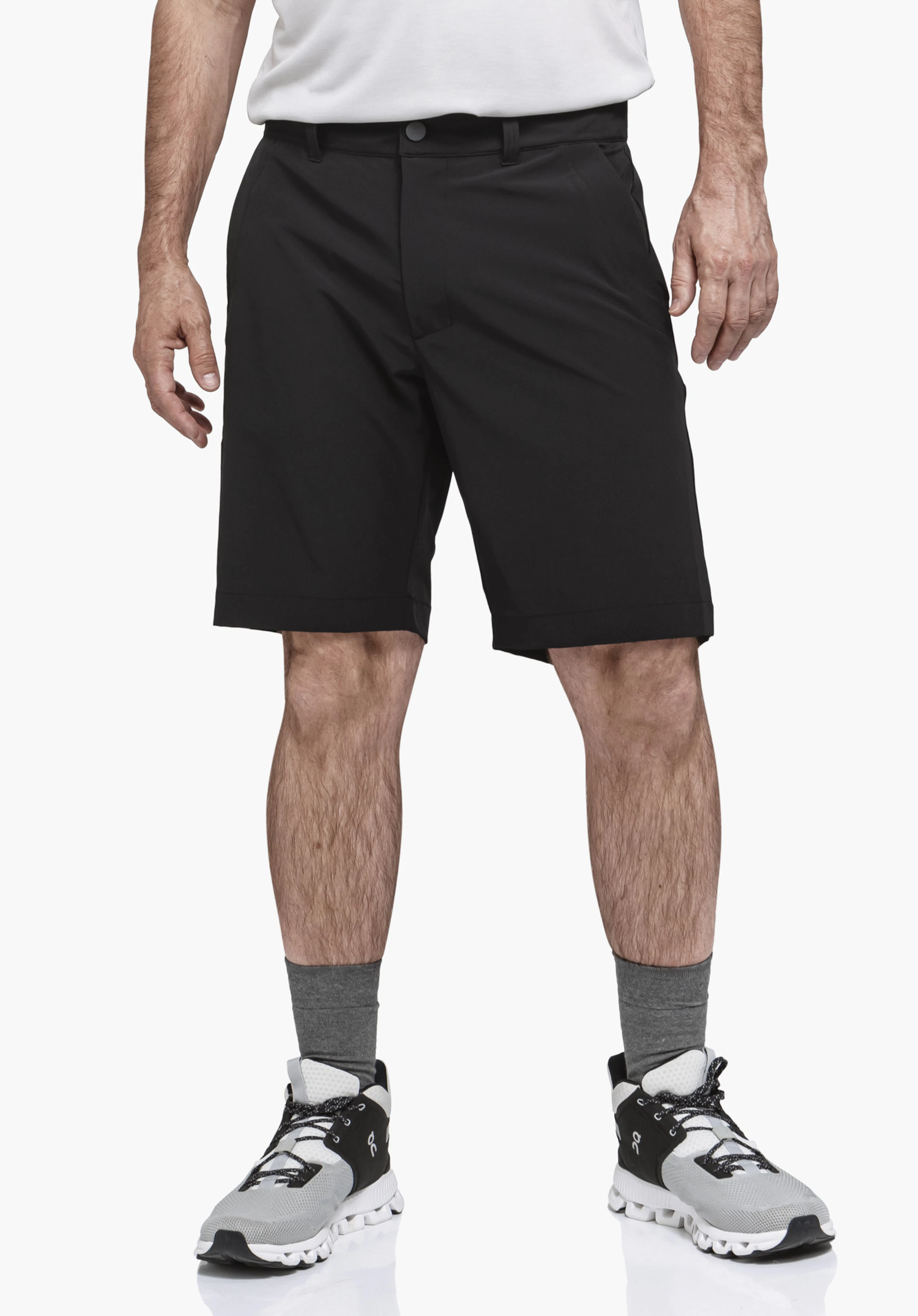 Shorts Style Gharra MNS. A model stands facing the front and presents the black shorts in a front view. The outfit is complemented by a light top, grey socks and trainers, photographed against a neutral background.