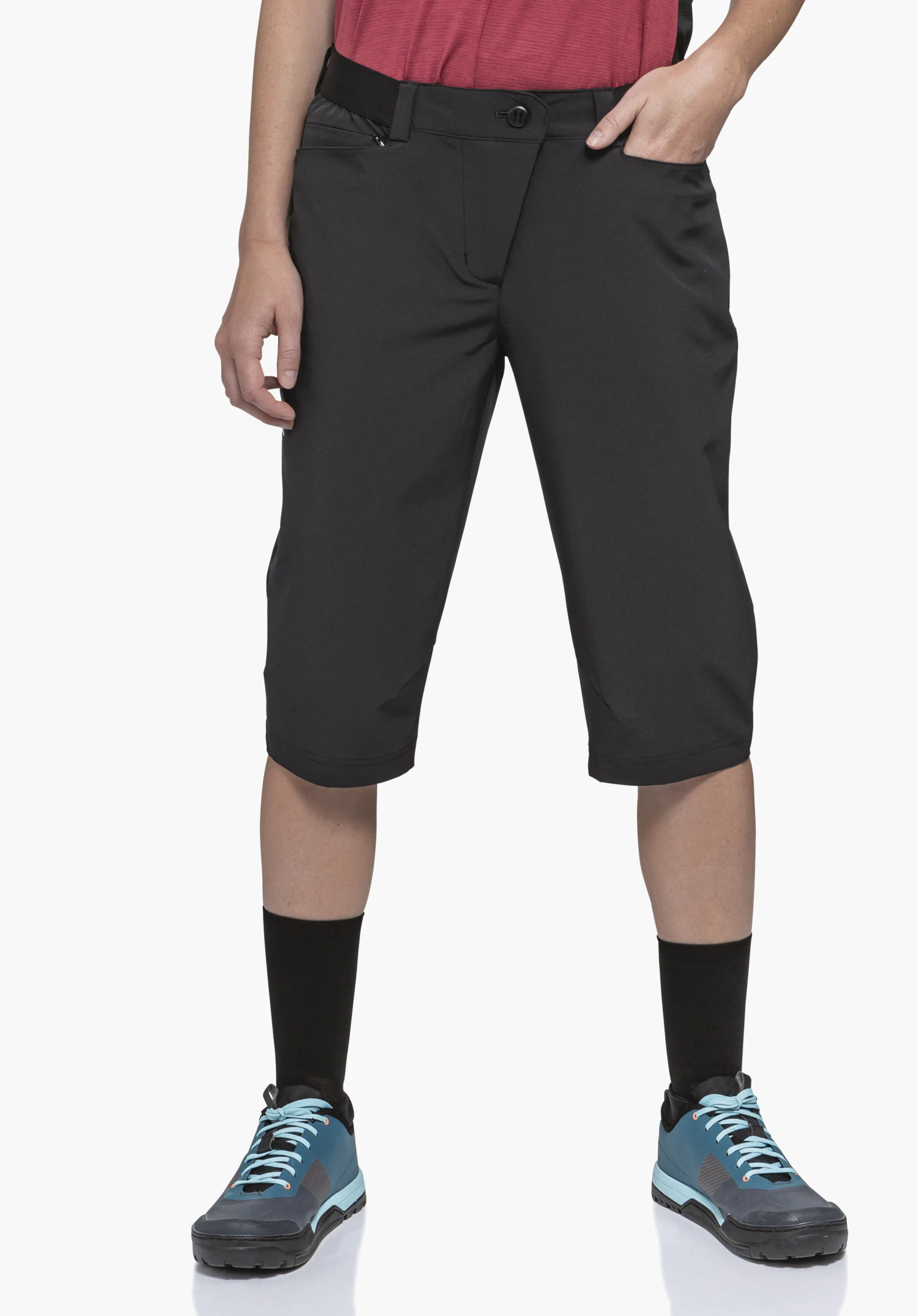 Capri Pants Style Keitele WMS women’s black 3/4-length trousers; worn by a standing person with right hand in pocket and left hand relaxed; red shirt, light blue shoes, black socks; white background.