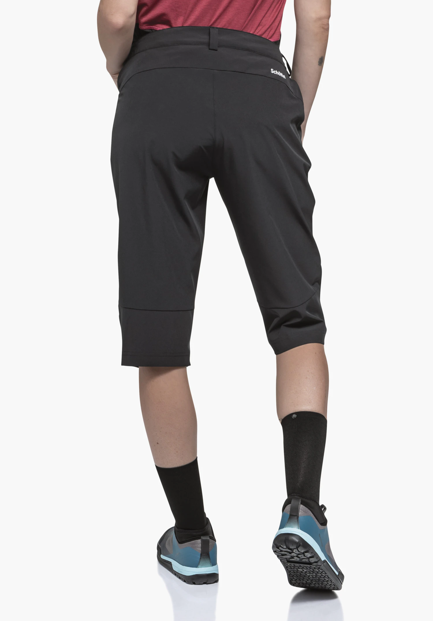 Capri Pants Style Keitele WMS Schwarze knielange Shorts, Person steht rückwärts mit Händen in Taschen, rotes Oberteil, schwarze Socken, hellblau-schwarze Schuhe vor weißem Hintergrund. Text: "Schöffel"