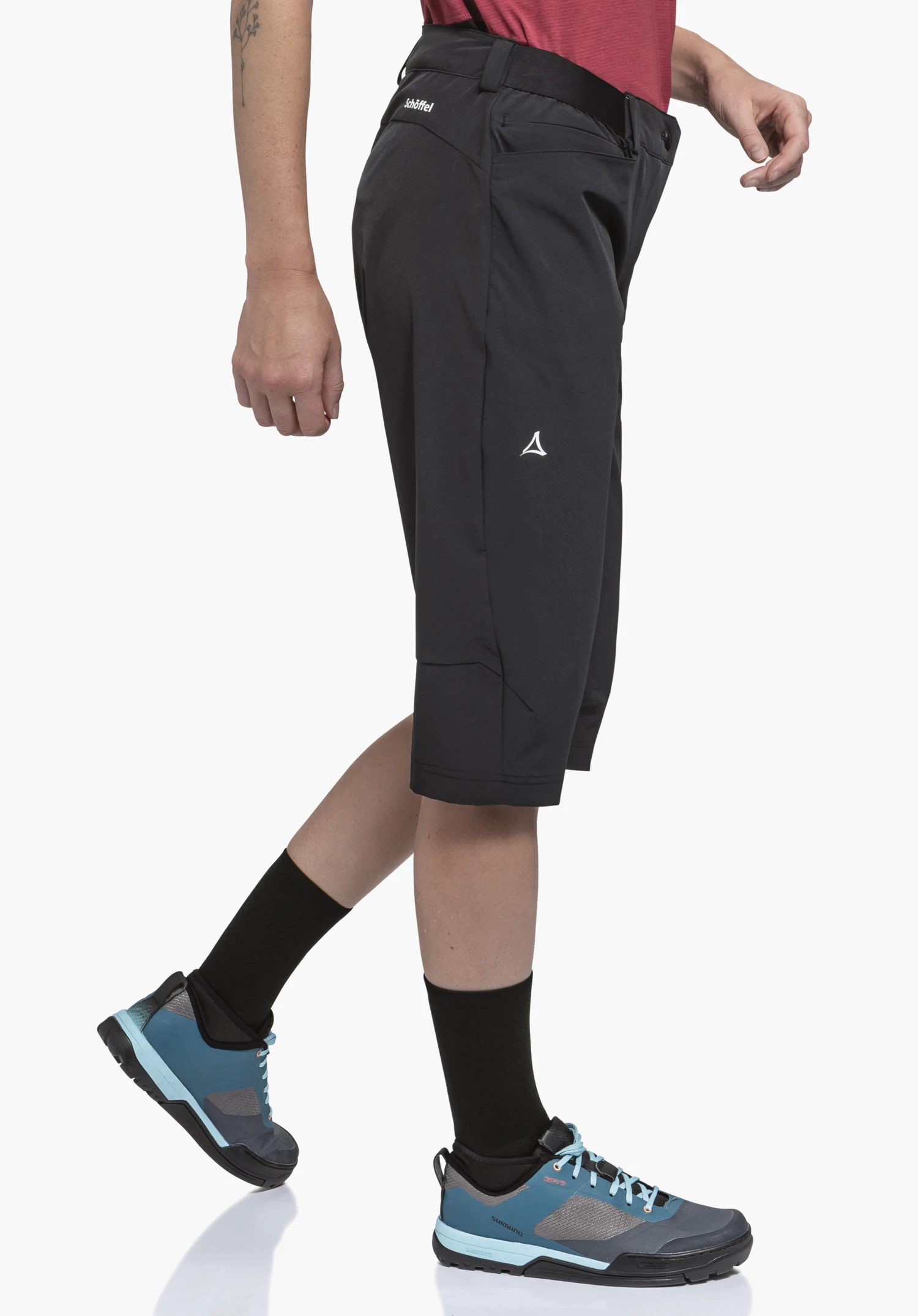 Capri Pants Style Keitele WMS Dunkelgraue knielange Shorts (Schöffel-Logo, Text: 'Schöffel') werden von einer gehenden Person getragen; rotes Oberteil, schwarze Socken, blaue Wanderschuhe, weißer Studiohintergrund.