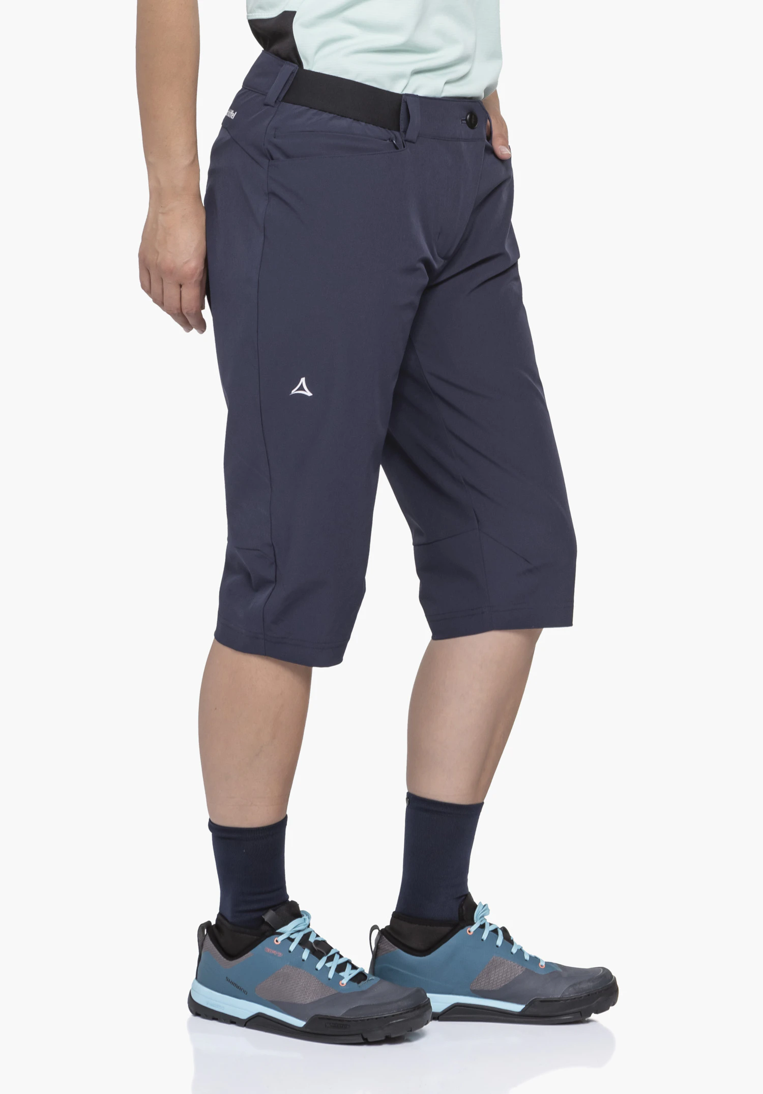 Capri Pants Style Keitele WMS in Blau. Das Model steht seitlich vor einem neutralen Hintergrund und präsentiert die Hose in ihrer funktionalen Passform. Auf dem Oberschenkel ist das dezente Schöffel-Logo gut zu erkennen.