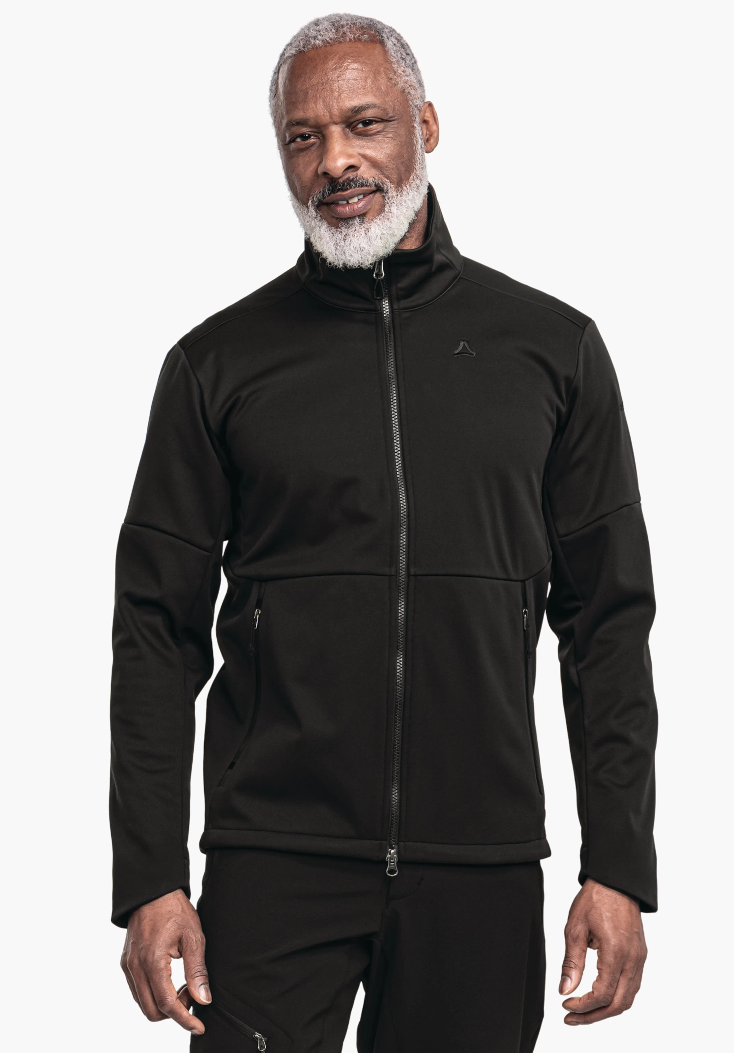 Softshell Jk Style Fracon MNS schwarze Reißverschlussjacke: Person steht frontal, trägt Jacke mit Schöffel-Logo auf der Brust, leichtes Lächeln, weißer Bart, entspannte Haltung, neutraler weißer Studiohintergrund.