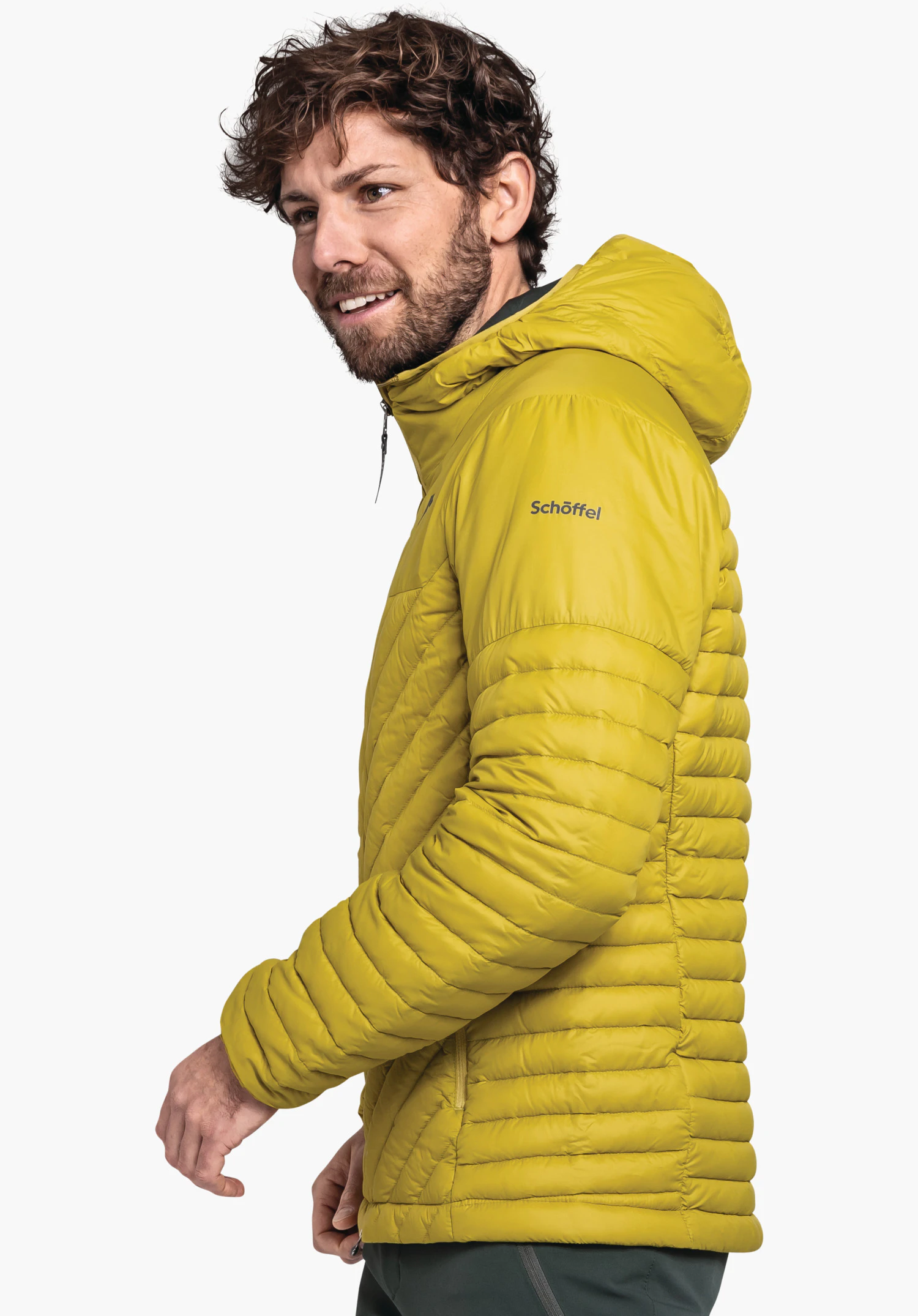 Down Jacket Style Cascata MNS veste matelassée jaune clair portée par un homme barbu souriant regardant de profil; veste avec capuche, fermeture éclair et inscription "Schöffel". Fond blanc uni.
