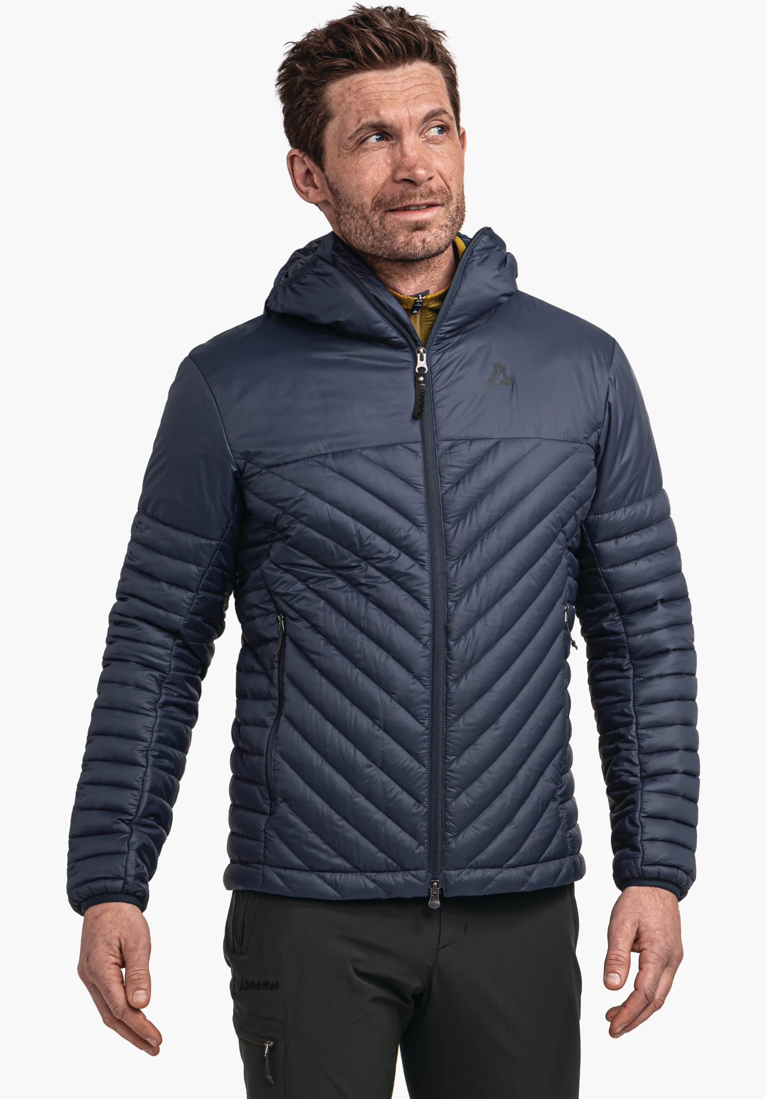 Down Jacket Style Cascata MNS Dunkel blaue Steppjacke, getragen von einem Mann, der nach oben rechts blickt; Schöffel-Logo auf der Brust; Text "Schöffel" auf der dunkel grünen Hose; weißer Studiohintergrund.