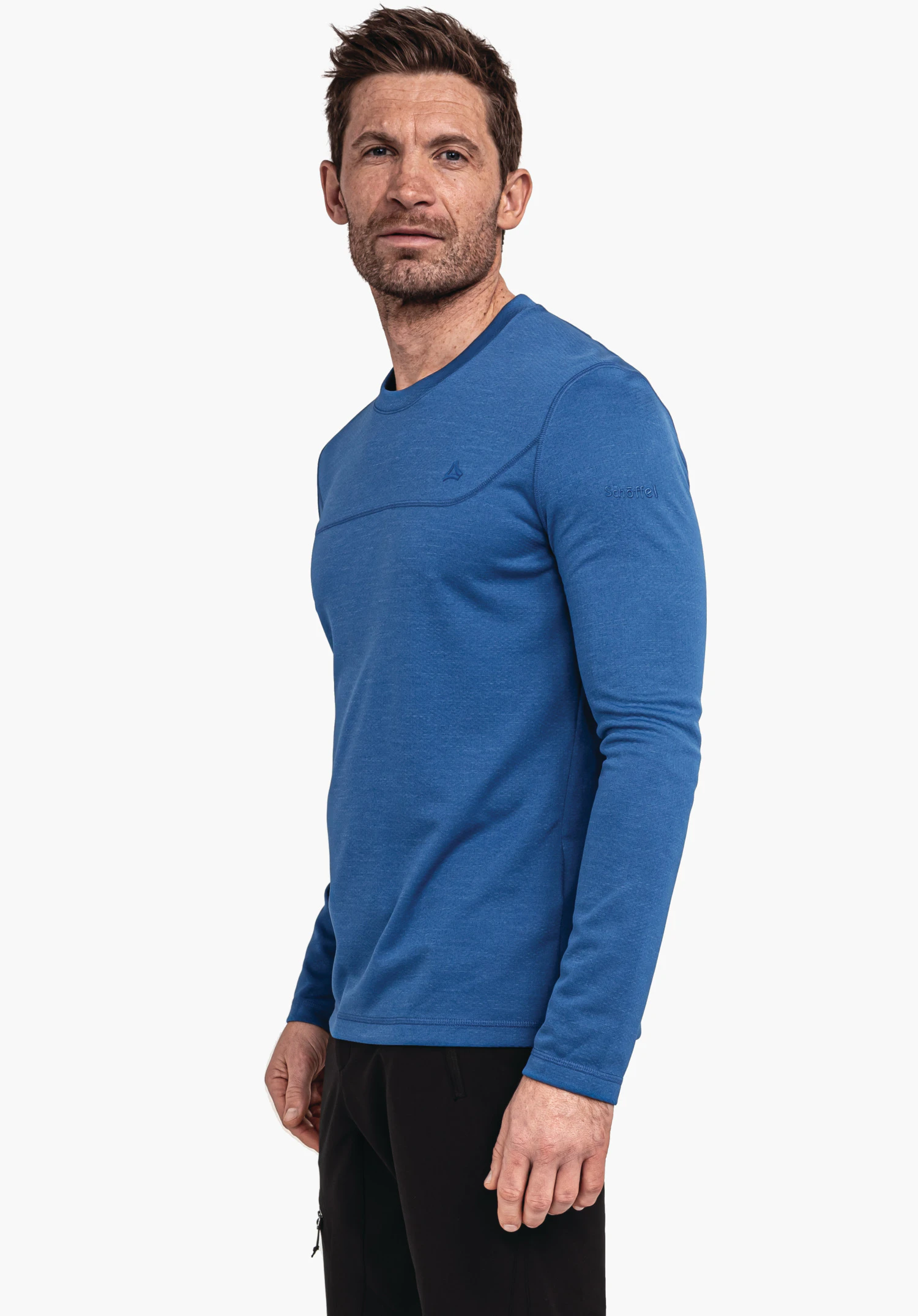 CIRC Longsleeve Style Smue MNS Mann trägt blaues Langarmshirt mit kleinem Schöffel-Logo auf der Brust und gesticktem Schriftzug "Schöffel" am Ärmel; steht seitlich, Arme entspannt, vor weißem Hintergrund.