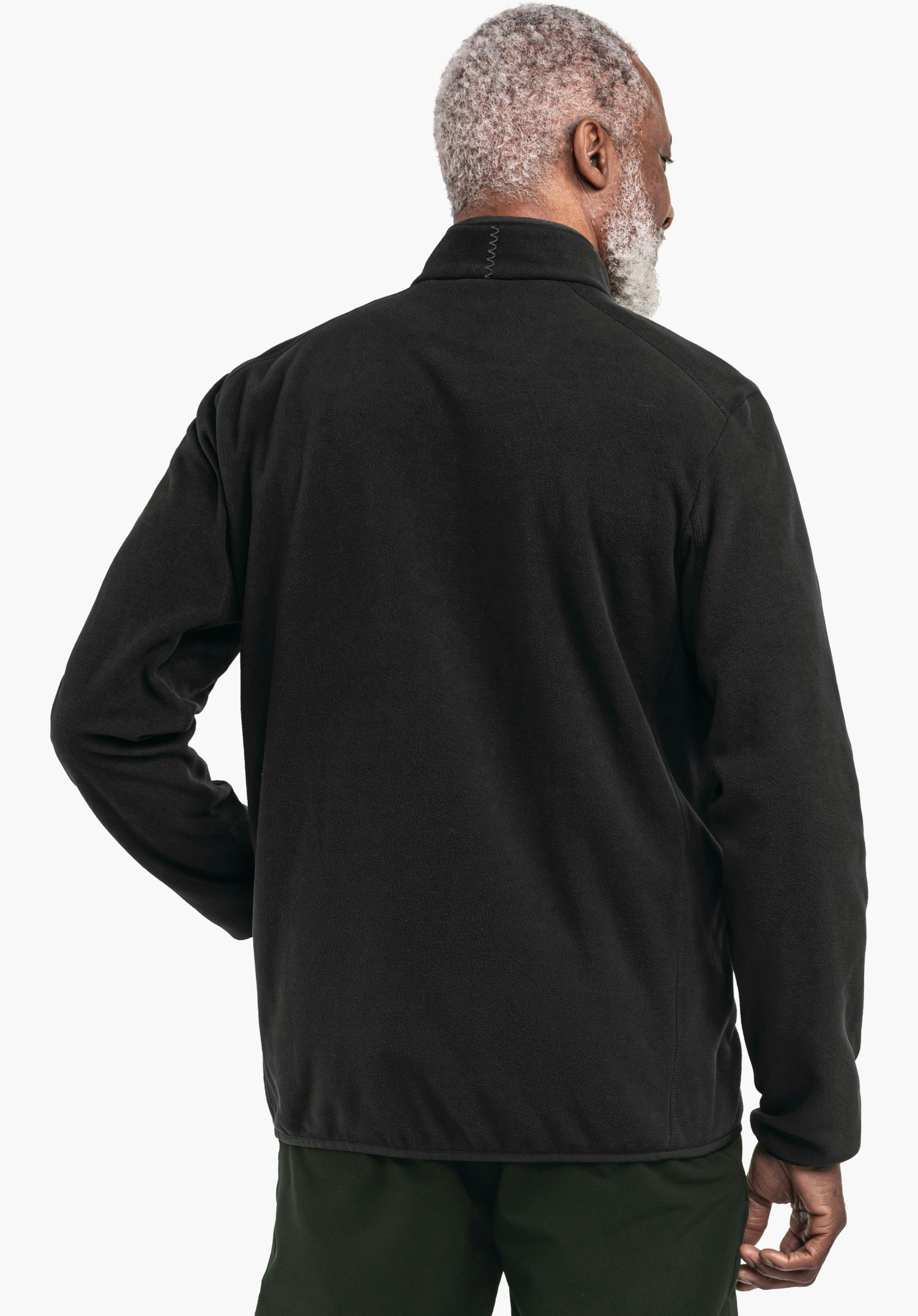 Fleece Jk Style Ash MNS Schwarze Fleecejacke an einem Mann, der rückwärts steht und leicht nach rechts blickt; hellgraues Haar; Umgebung neutral hell, Hose dunkelgrün.