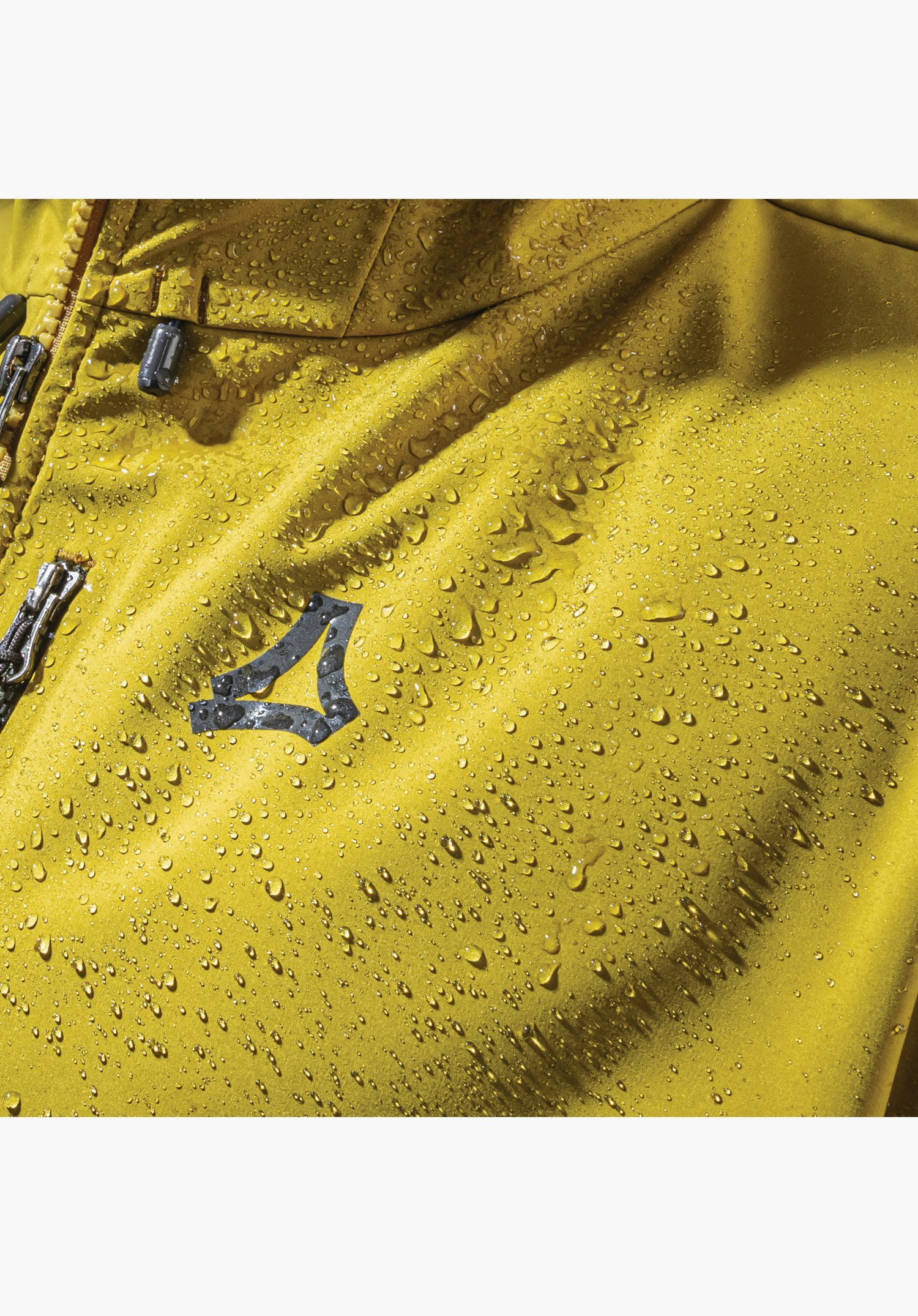 Softshell Jk Style Milagle MNS zeigt die wasserabweisende Eigenschaft des gelben Materials mit abperlenden Wassertropfen und das Schöffel-Logo. Nahaufnahme vor weißem/neutralem Hintergrund.