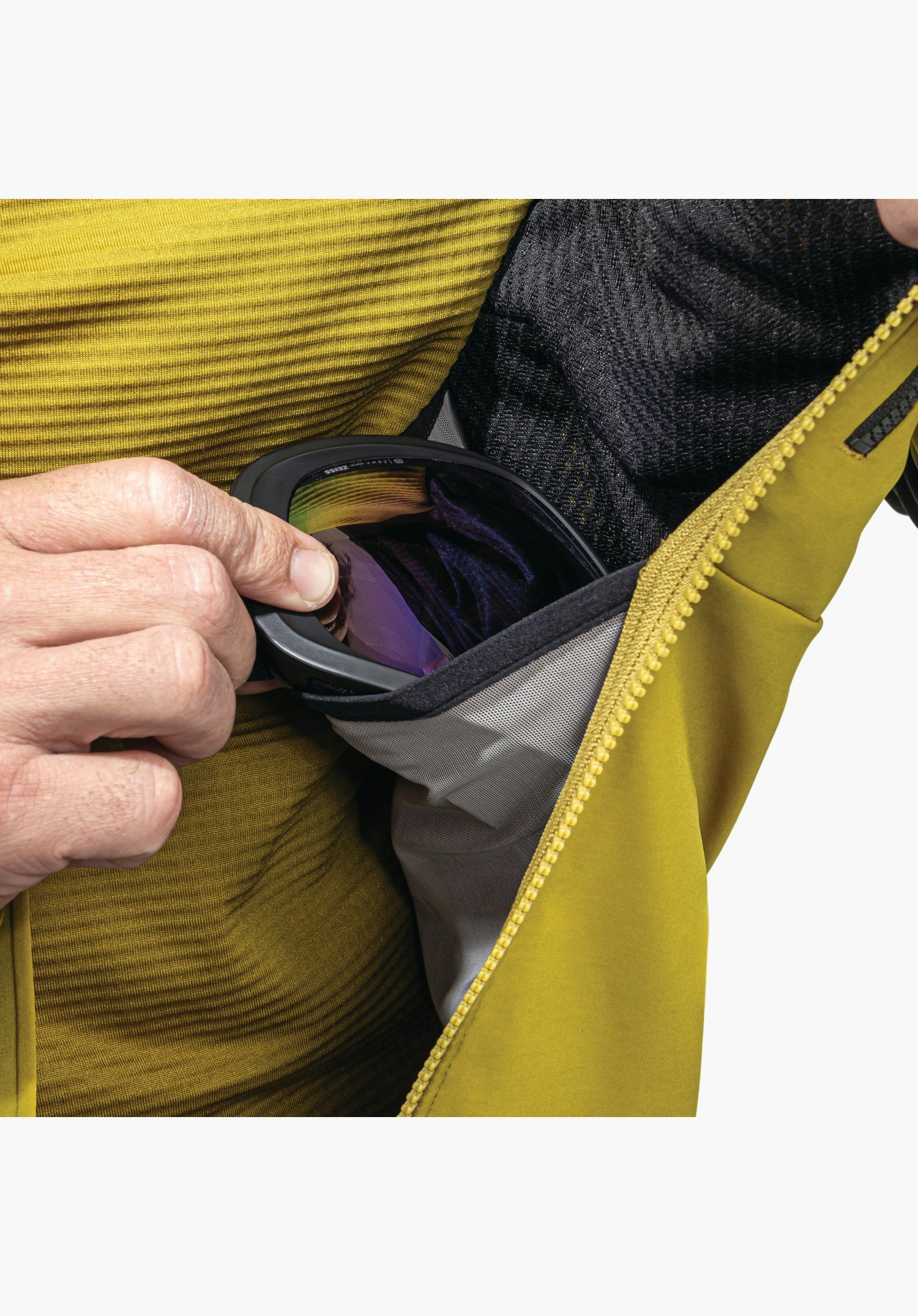 Softshell Jk Style Milagle MNS, Innenseite mit geräumiger Mesh-Tasche, in die gerade eine Skibrille gesteckt wird, sowie der gelbe Reißverschluss, Nahaufnahme vor weißem/neutralem Hintergrund.