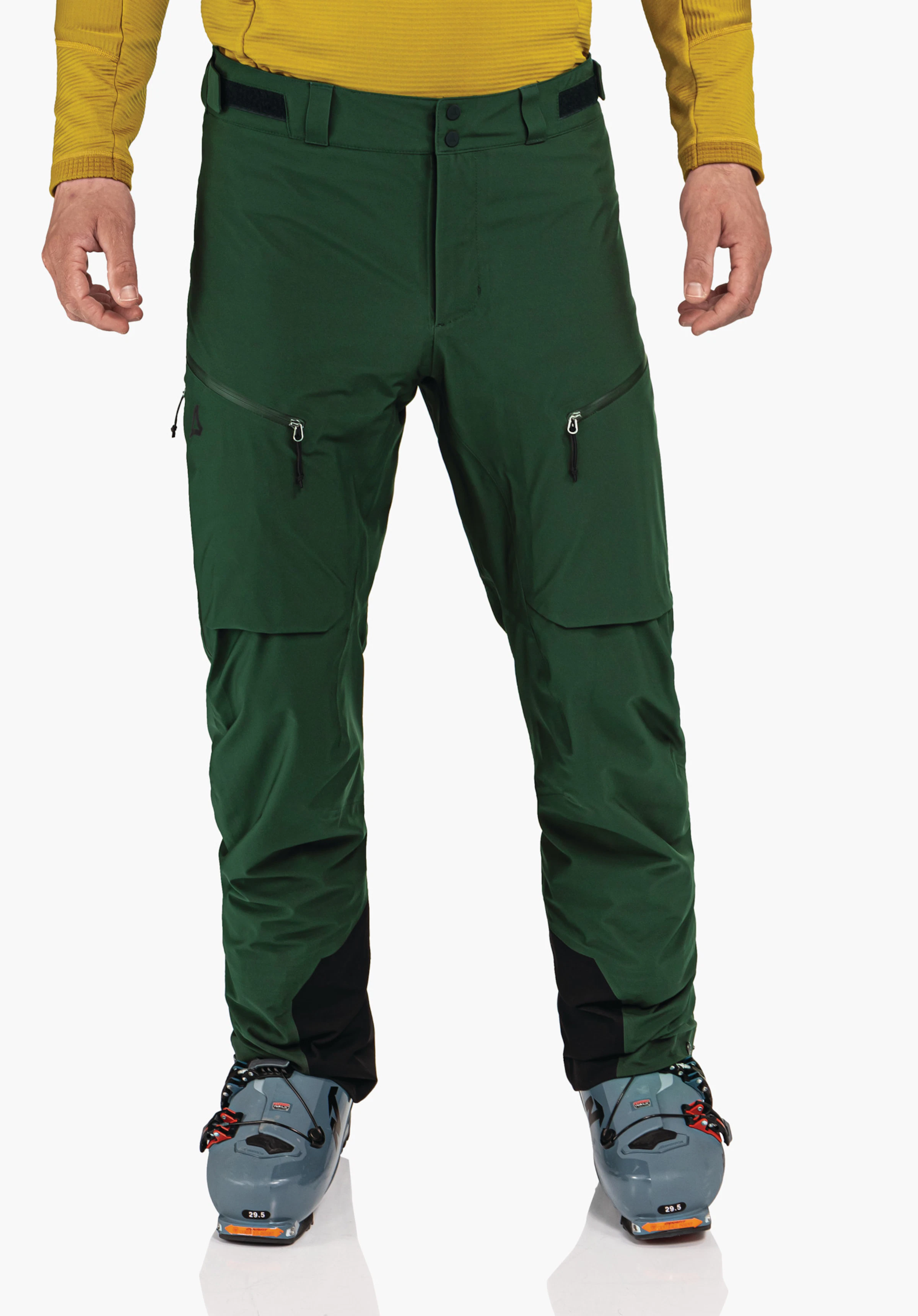 Pants Style Milagle MNS dunkelgrüne Outdoor-Hose mit Schöffel-Logo, wird von einer stehenden Person getragen; Kontext: gelbes Oberteil, hell blau Skischuhe auf weißem Hintergrund. Text: 29.5