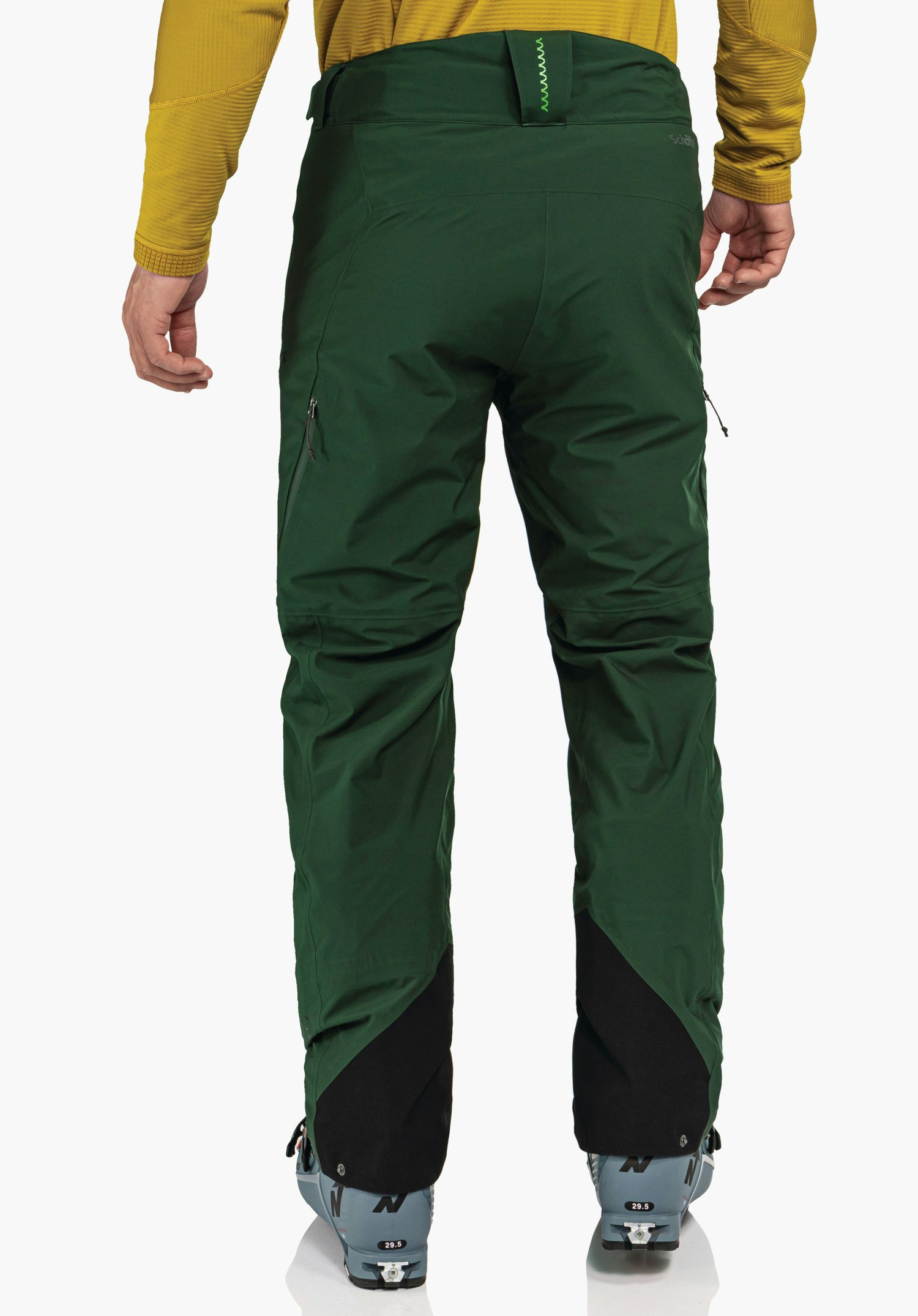 Pants Style Milagle MNS dark green ski trousers worn from the back, showing zip pockets and black leg reinforcements; person stands wearing a light yellow top and light blue ski boots against a white background.