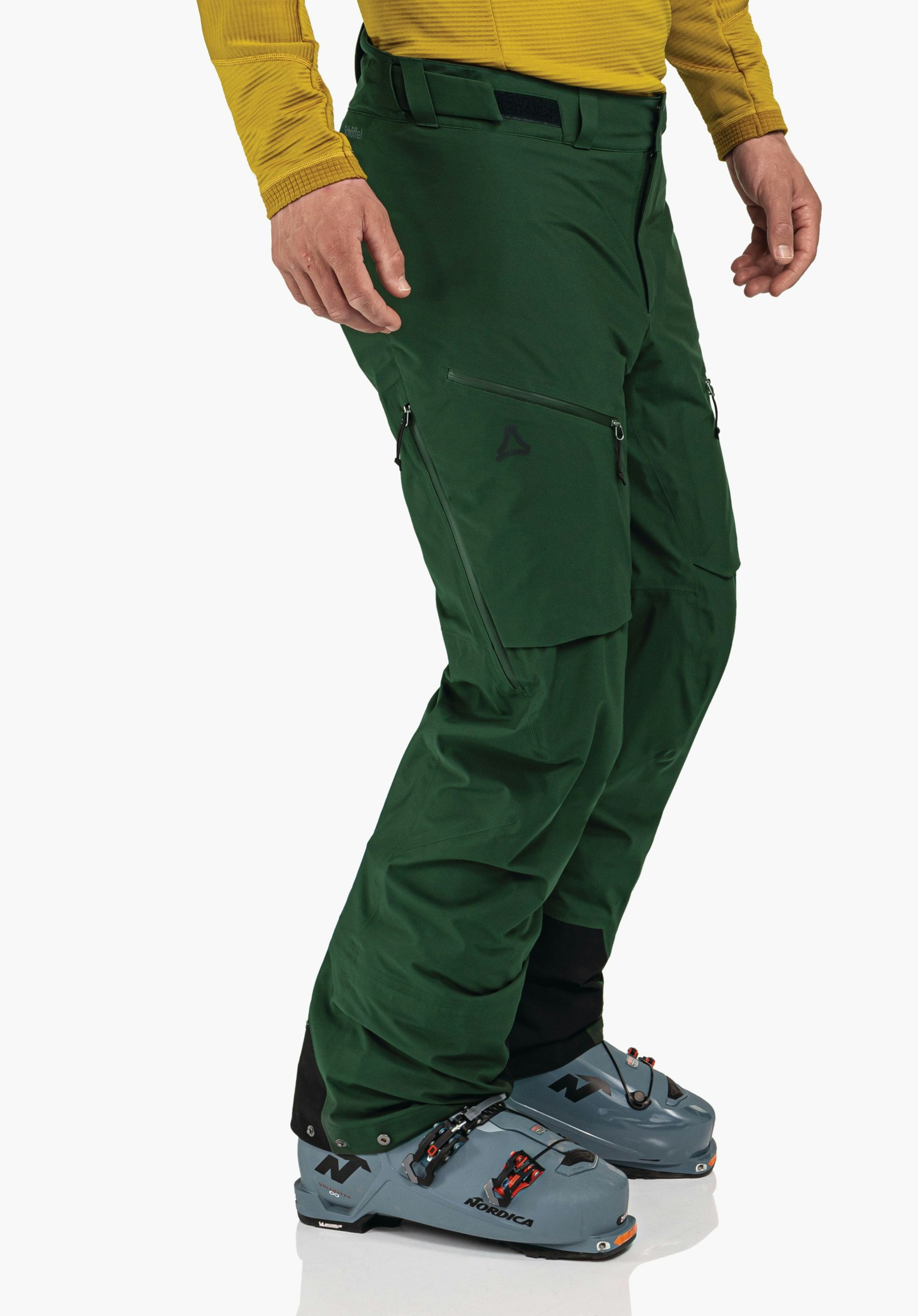 Pants Style Milagle MNS dark green ski trousers worn by a standing person slightly side-on, hands relaxed at sides; light yellow top, light blue ski boots on white studio background. Schöffel logo on thigh. Text: "NORDICA".