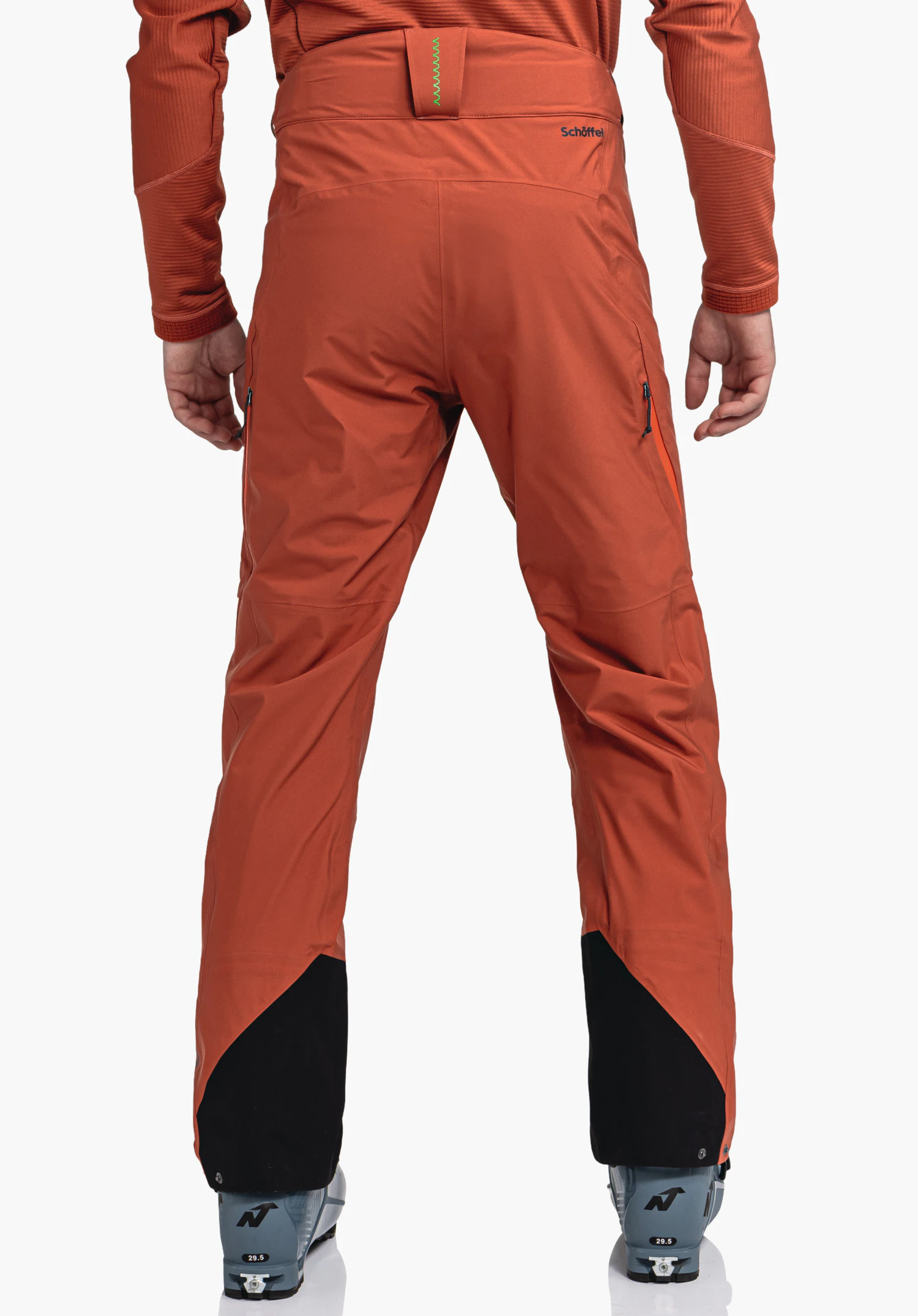 Pants Style Milagle MNS dark orange ski trousers; person stands with back to the camera, legs slightly apart, black leg reinforcements, ski boots visible. Text: “Schöffel”, “N”, “29.5”.