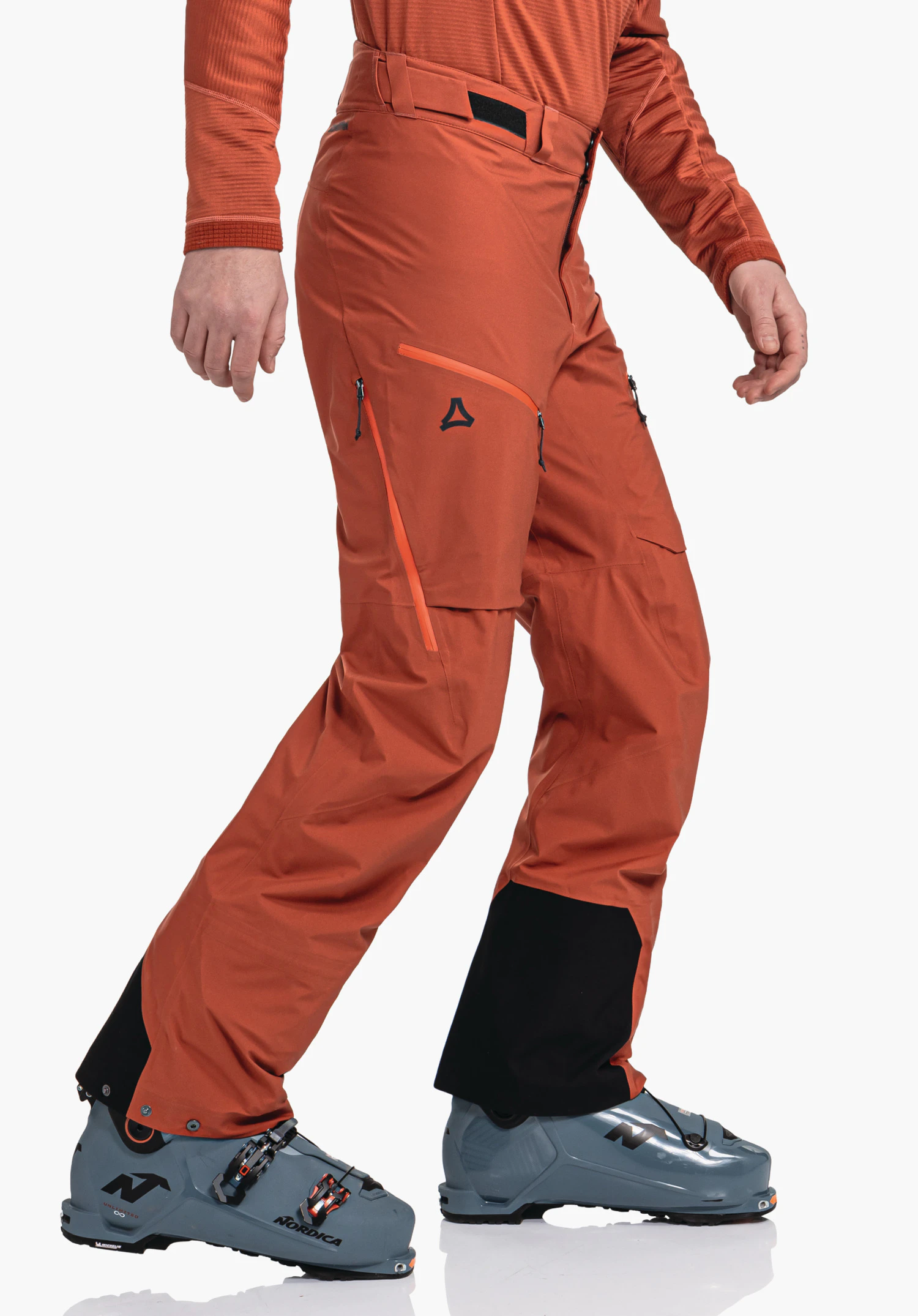 Pants Style Milagle MNS dark orange ski trousers with Schöffel logo on the outer thigh, person takes a step; light blue ski boots with lettering "NORDICA", white studio background.