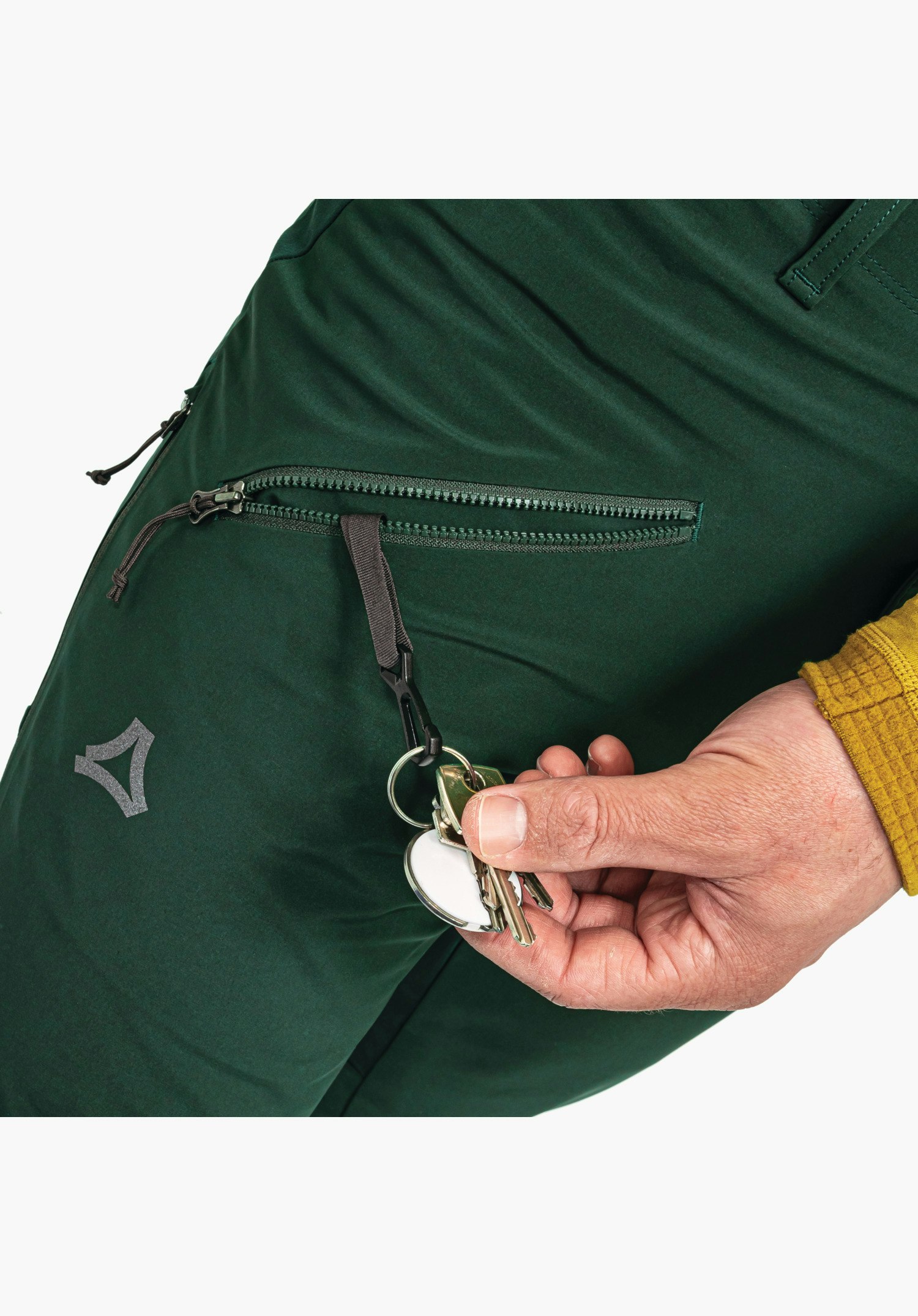 Softshell Pt Style Milagle MNS montre la poche zippée ouverte sur le tissu softshell vert avec un porte-clés intégré retenant des clés, ainsi que le logo Schöffel en gros plan sur fond blanc.