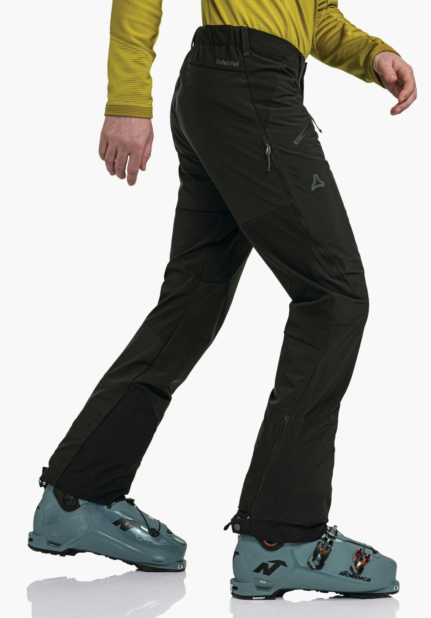 Softshell Pt Style Milagle MNS schwarze Hose, gehende Person macht Schritt vorwärts, dunkelgelbes Oberteil, dunkelblaue Skischuhe, weißer Studiohintergrund; Schöffel-Logo sichtbar. Text im Bild: "Schöffel", "Nordica".