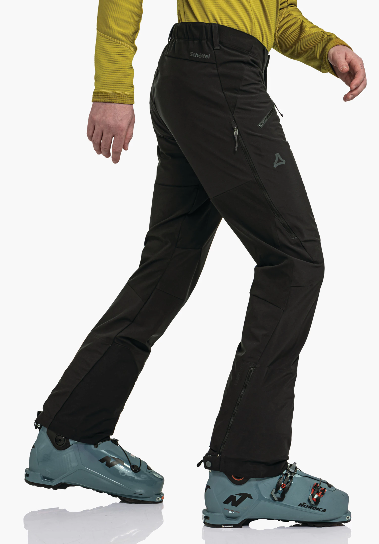Softshell Pt Style Milagle MNS black trousers worn by a walking person stepping forward, dark yellow top, dark blue ski boots, white studio background; Schöffel logo visible. Text in image: "Schöffel", "Nordica".