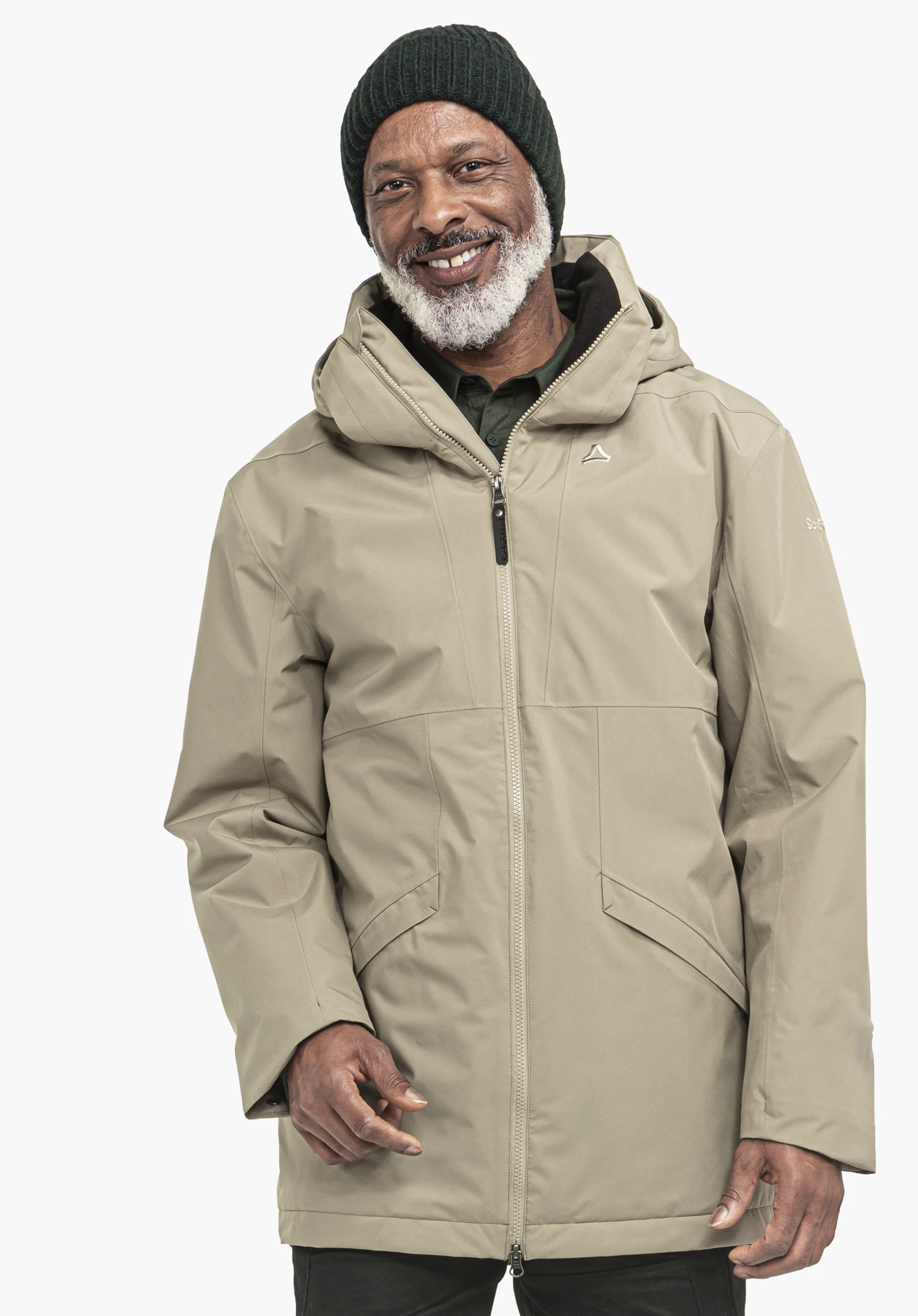 Ins Parka Style Malkay MNS homme portant un parka marron clair avec logo Schöffel, souriant, debout devant un fond clair et neutre; bonnet vert foncé, barbe blanche et pantalon noir.