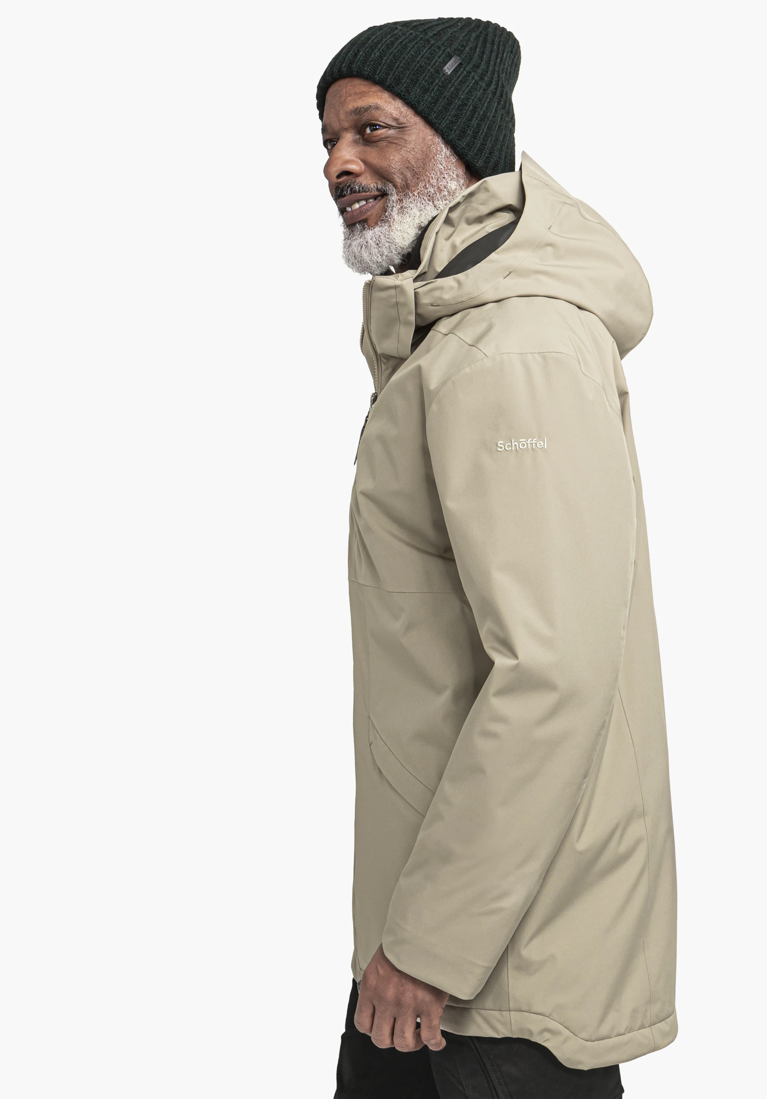 Ins Parka Style Malkay MNS homme portant une veste à capuche avec bonnet vert foncé, debout de profil et souriant; fond blanc neutre. Texte sur la manche: "Schöffel".