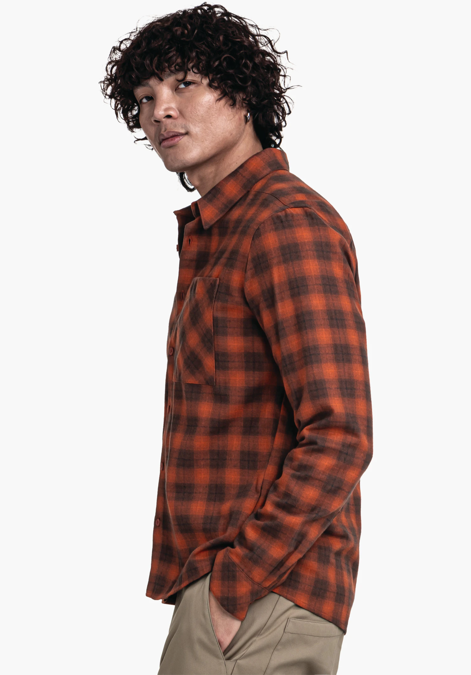 Shirt Style Kleion MNS man stands side-on with hand in pocket, wears an orange checked shirt and beige trousers in front of a white background; curly dark hair, looks towards the camera.