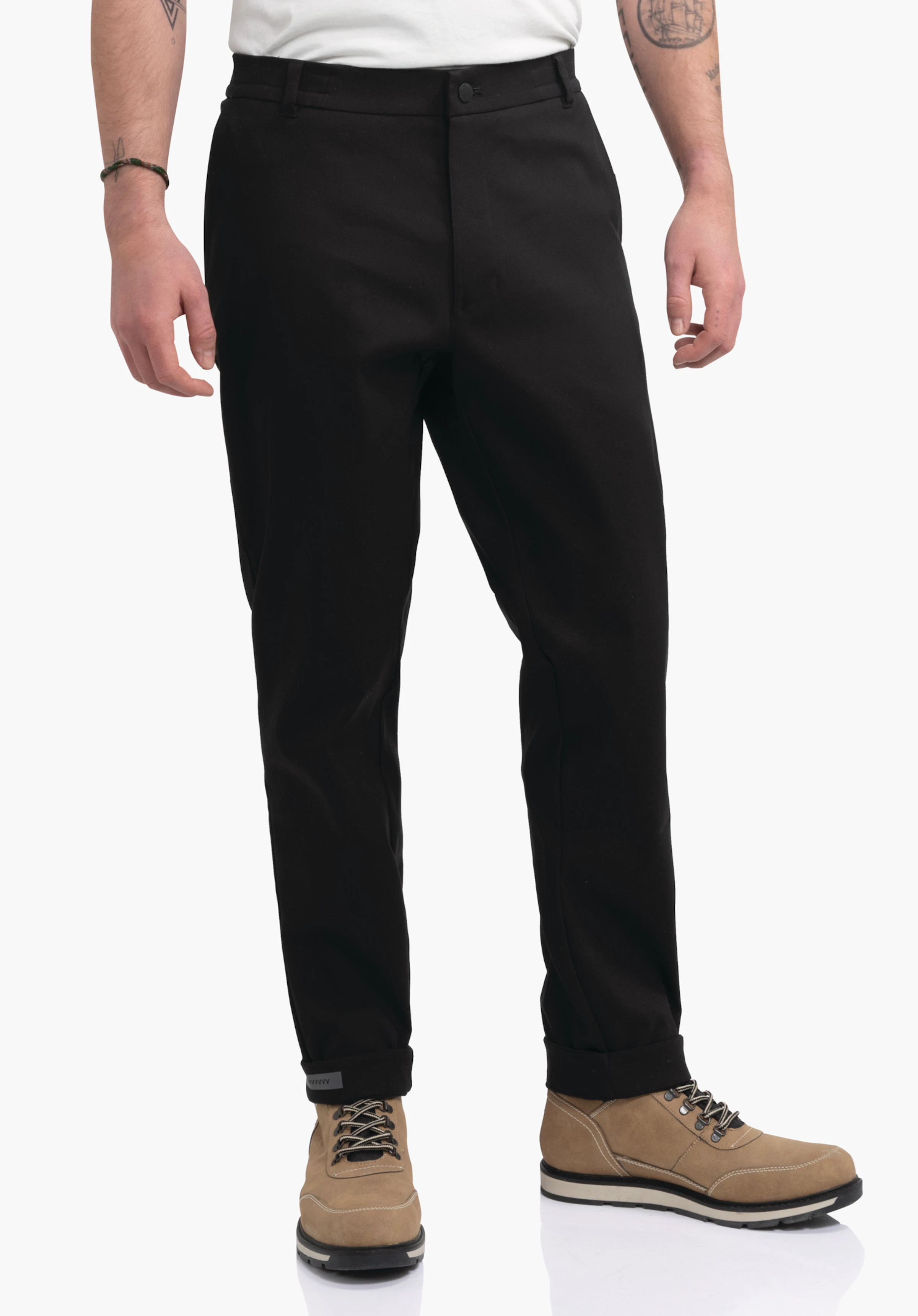Pants Style Zourga MNS black trousers worn by a standing person, legs slightly rolled up; context: light background, visible tattooed forearms, leather lace-up boots.