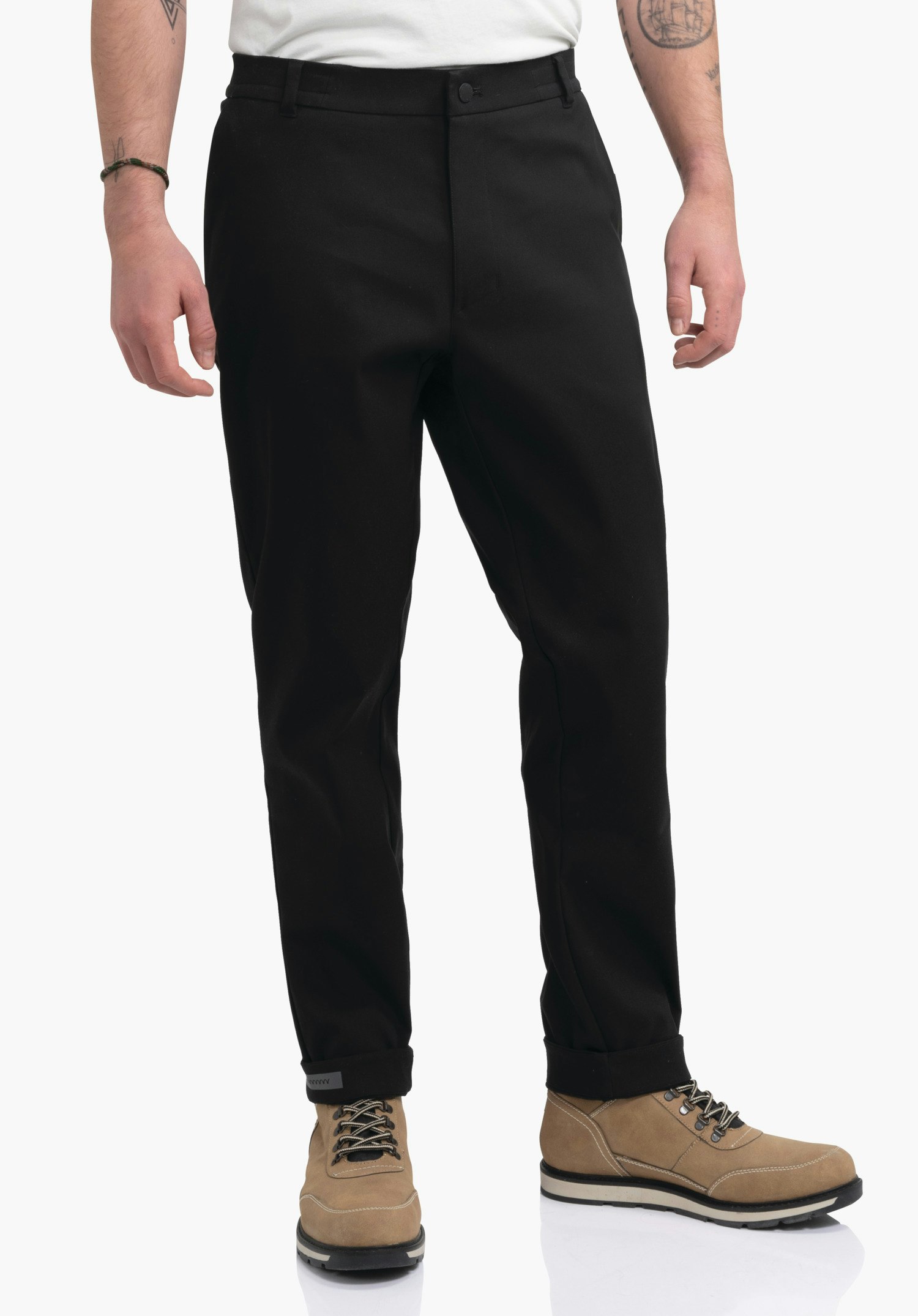 Pants Style Zourga MNS schwarze Hose, getragen von stehender Person, Beine leicht gekrempelt; Kontext: heller Hintergrund, sichtbare Unterarme mit Tattoos, lederne Schnürstiefel.