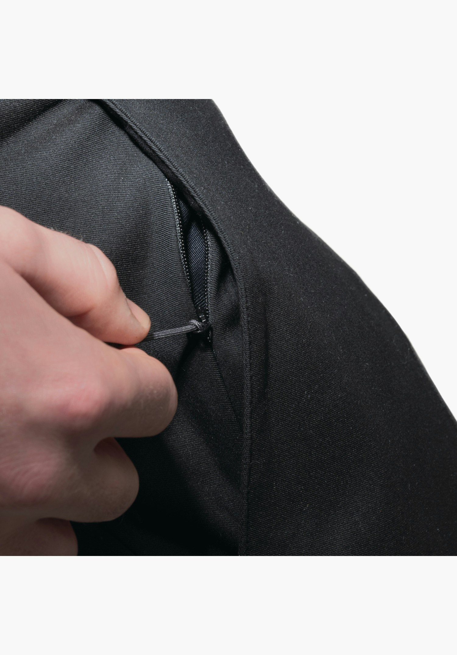Pants Style Zourga MNS. Detailansicht einer schwarzen Reißverschlusstasche, an deren Zipper-Puller eine Hand zieht. Nahaufnahme vor weißem Hintergrund.