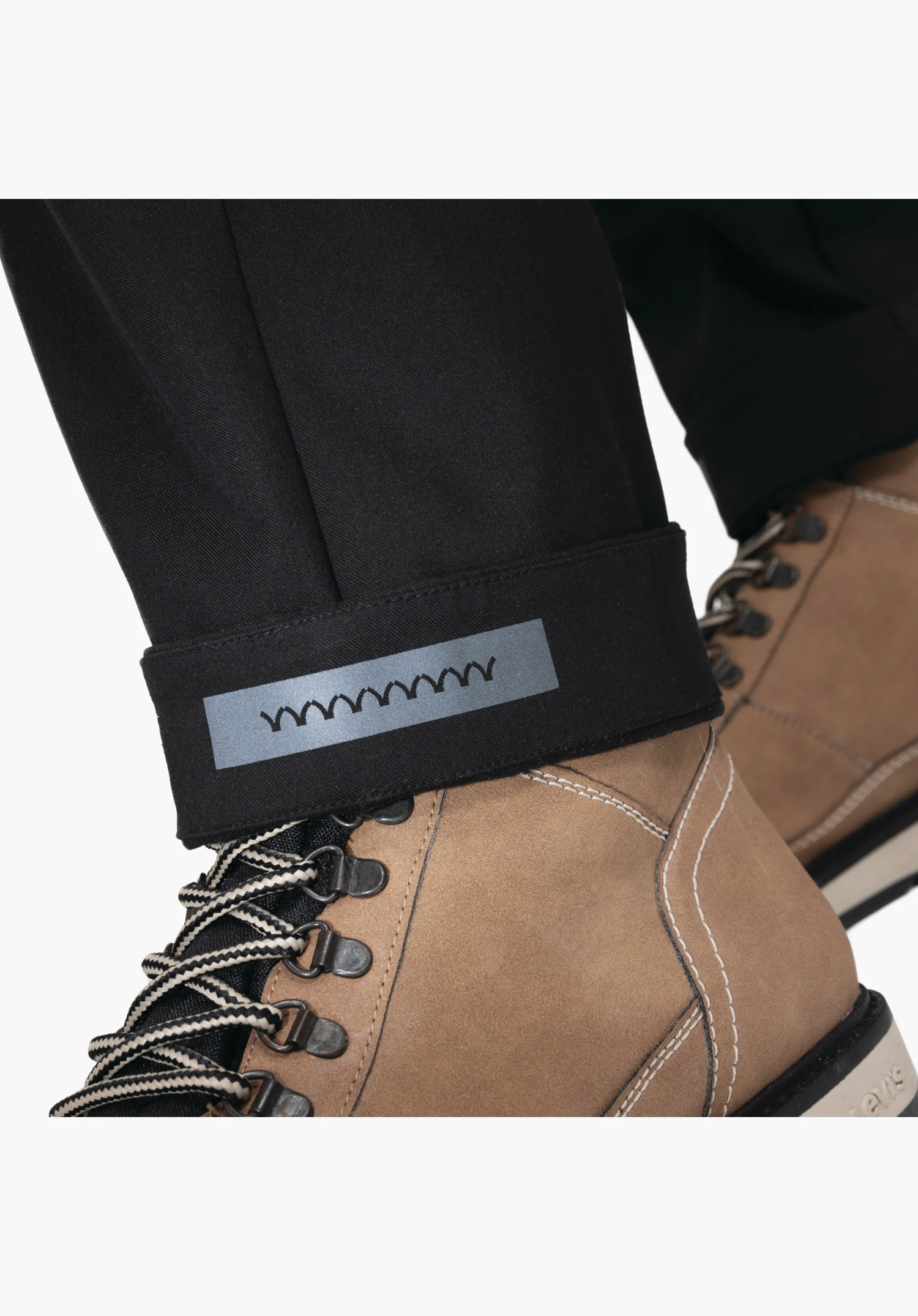 Pants Style Zourga MNS shows the turned-up black trouser hem featuring an integrated reflective strip. Close-up against a white background., Pants Style Zourga MNS shows the turned-up black trouser hem featuring an integrated reflective strip. Close-up against a white background.