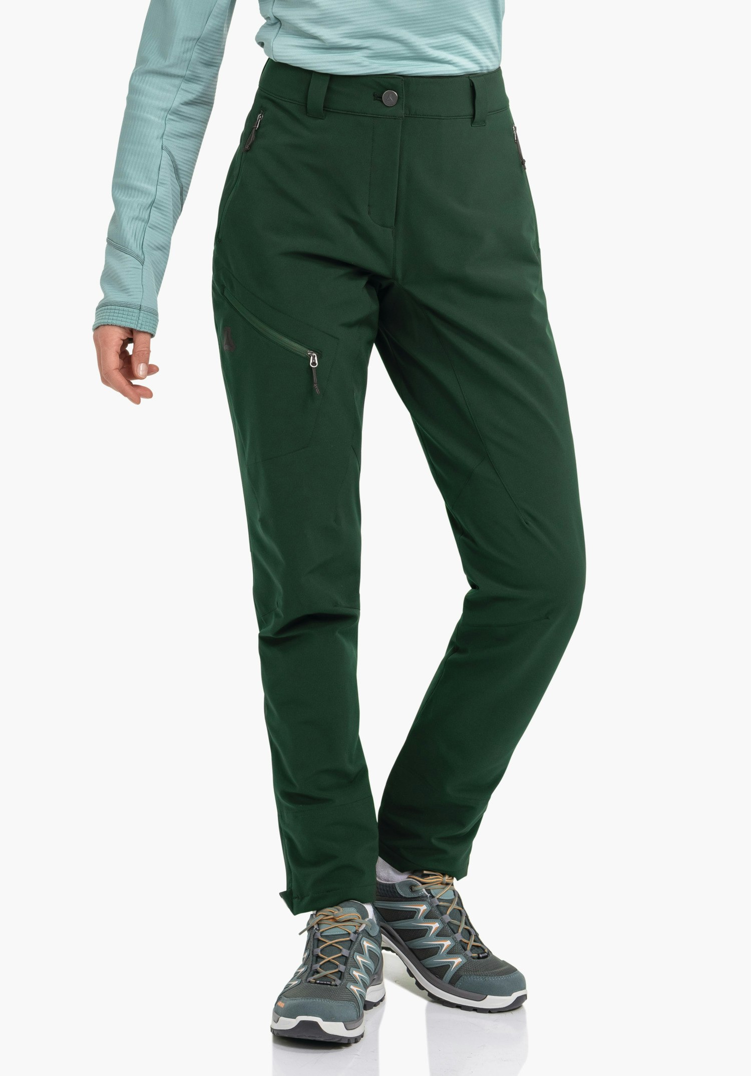 Pants Style Cascata WMS dark green trekking trousers, worn by a person in a light green top; legs slightly crossed, grey and blue hiking shoes; Schöffel logo on the outer left thigh; white studio background.