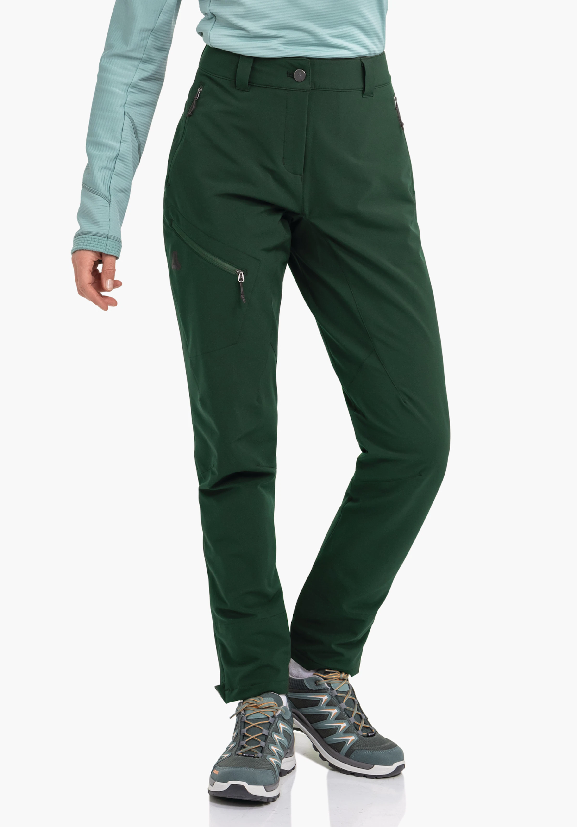 Pants Style Cascata WMS Pantalon de trekking vert foncé porté par une personne avec un haut vert clair; jambes légèrement croisées, chaussures de randonnée grises et bleues; logo Schöffel sur la cuisse extérieure gauche; fond studio blanc.