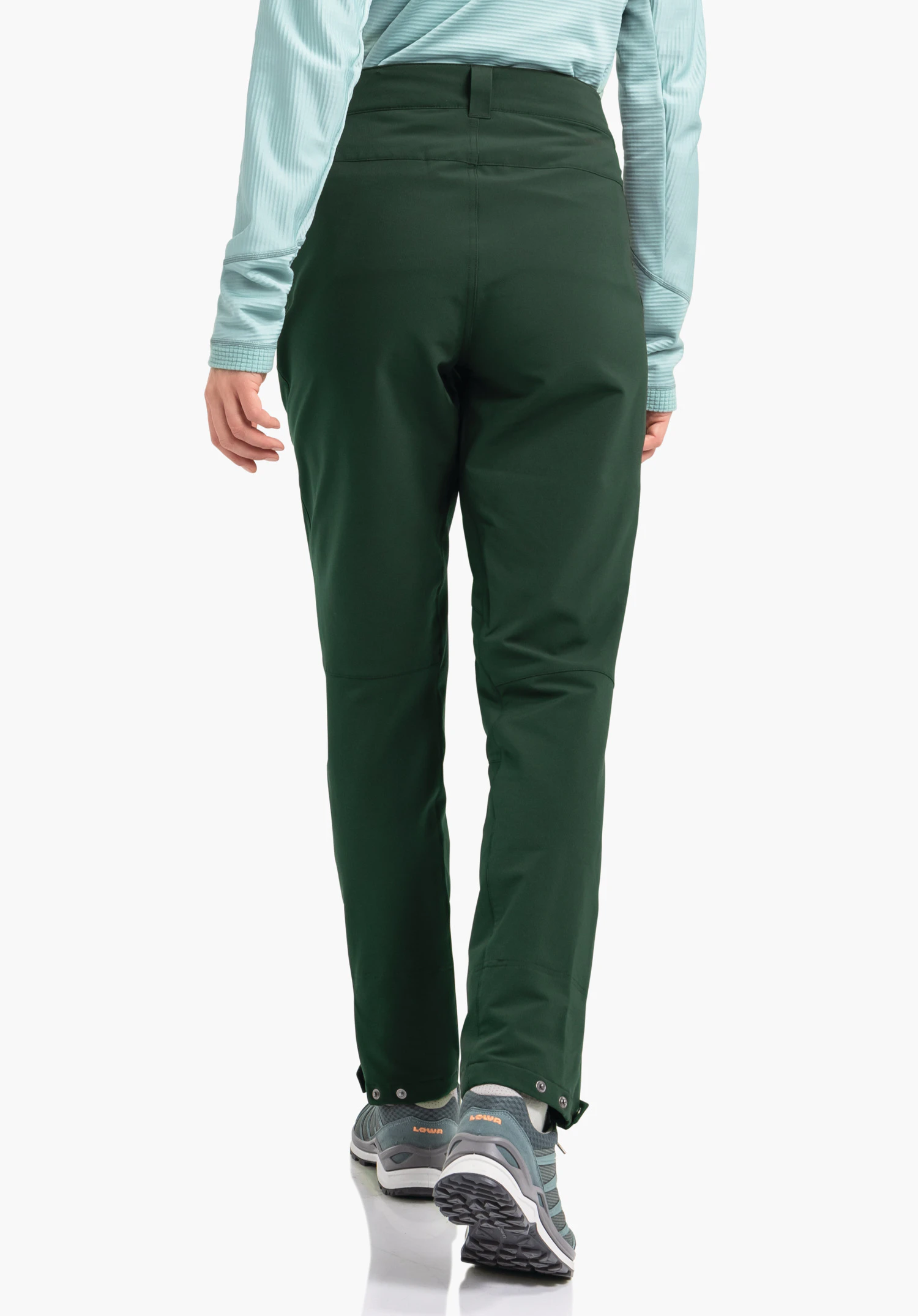 Pants Style Cascata WMS pantalon vert foncé, porté par une personne s’éloignant; contexte: fond studio blanc, haut manches longues bleu clair, chaussures de randonnée bleues avec texte orange « LOWA ».