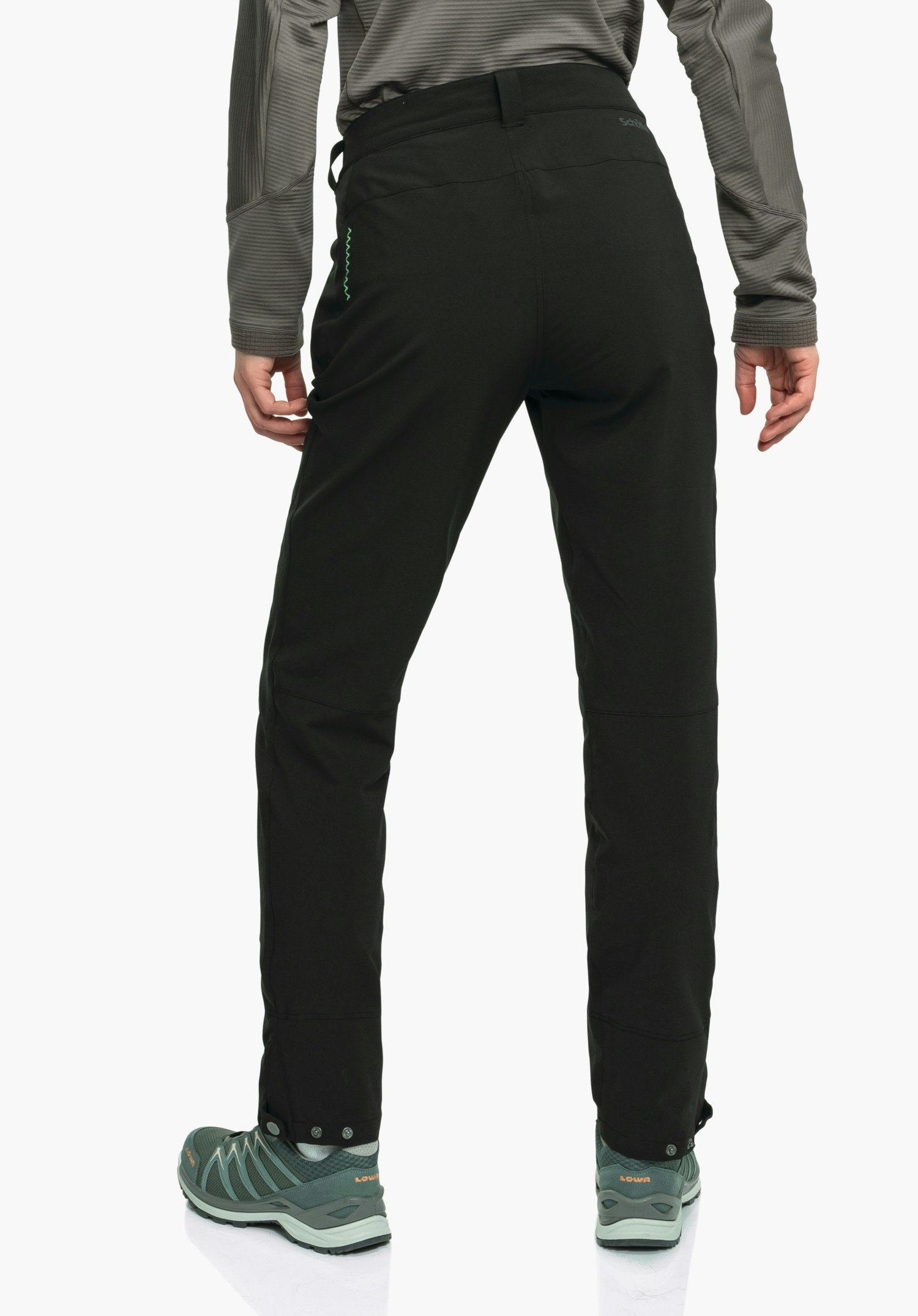 Pants Style Cascata WMS pantalon noir, porté de dos, posture debout jambes légèrement écartées; le modèle porte un haut manches longues gris et des chaussures de trekking vert foncé; fond studio blanc. Texte: « Schöffel », « LOWA ».