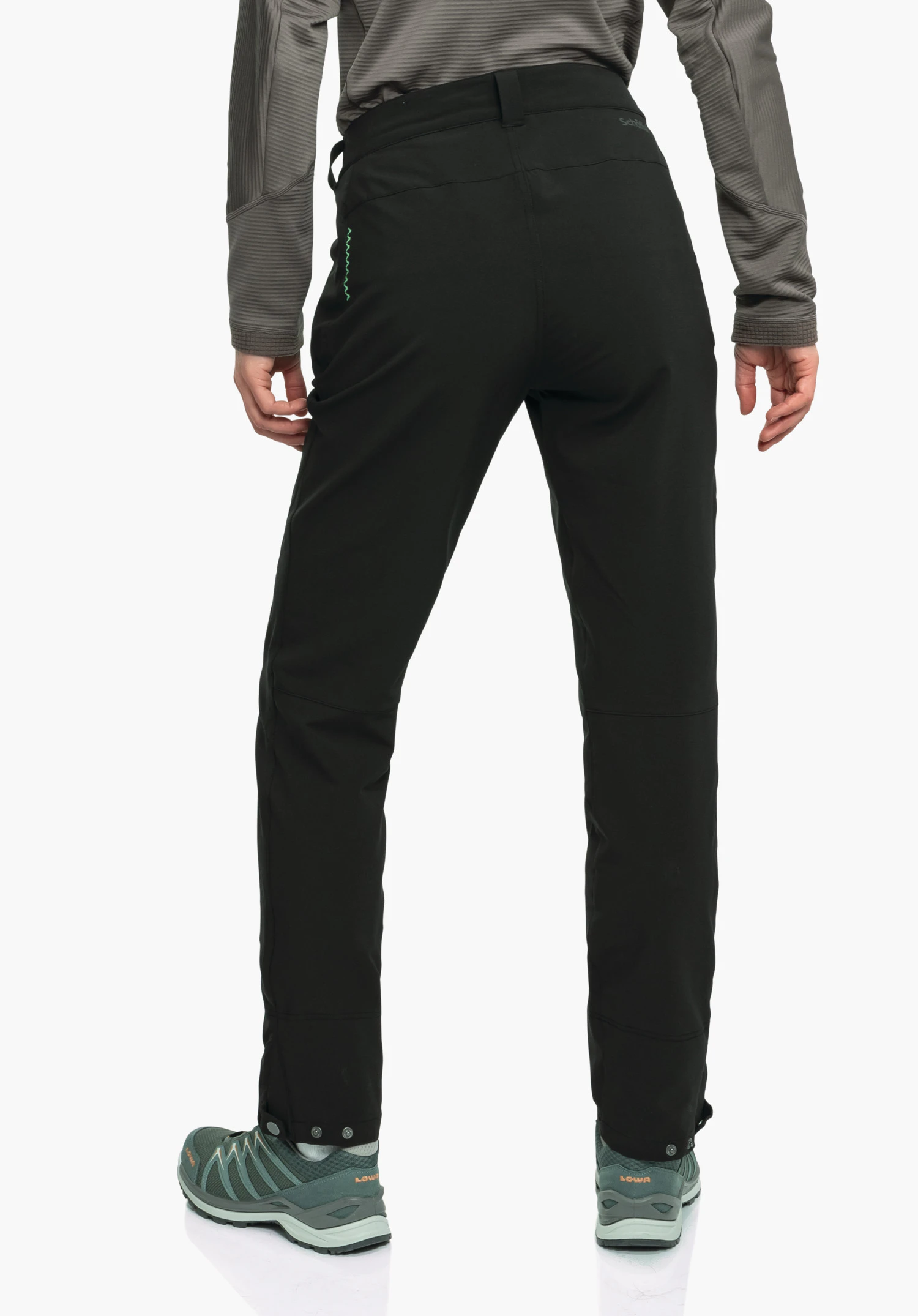 Pants Style Cascata WMS pantalon noir, porté de dos, posture debout jambes légèrement écartées; le modèle porte un haut manches longues gris et des chaussures de trekking vert foncé; fond studio blanc. Texte: « Schöffel », « LOWA ».