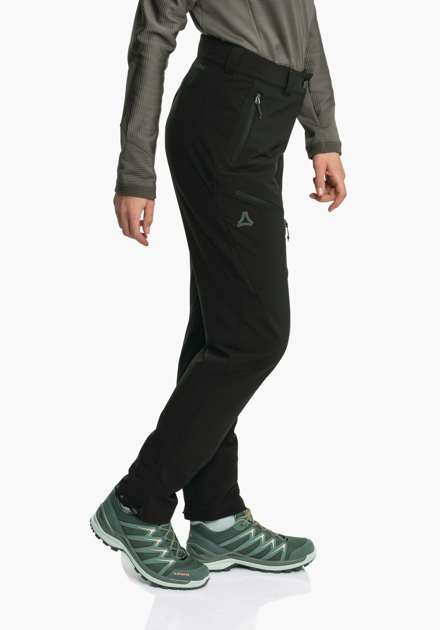 Pants Style Cascata WMS pantalon de randonnée noir avec logo Schöffel visible, poches zippées latérales; porté de profil, jambes croisées, avec chaussures de randonnée vertes portant l’inscription LOWA; fond studio blanc clair.