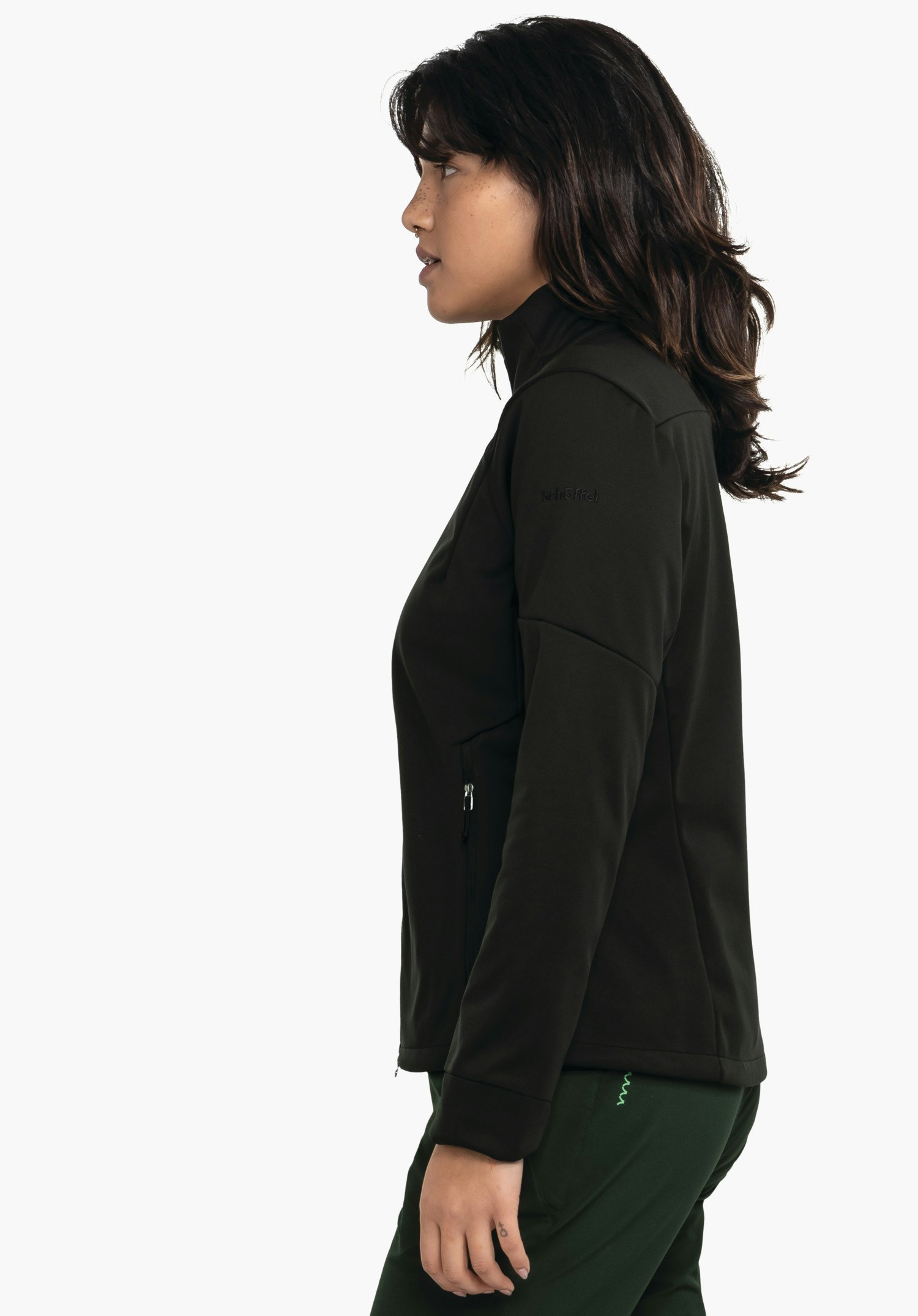 Softshell Jk Style Fracon WMS dunkel schwarze Jacke, getragen von Frau im Profil, steht seitlich, zeigt geschlossenen Reißverschluss und Tasche über dunkel grüner Hose, vor hellem weißem Hintergrund. Text: "Schöffel".
