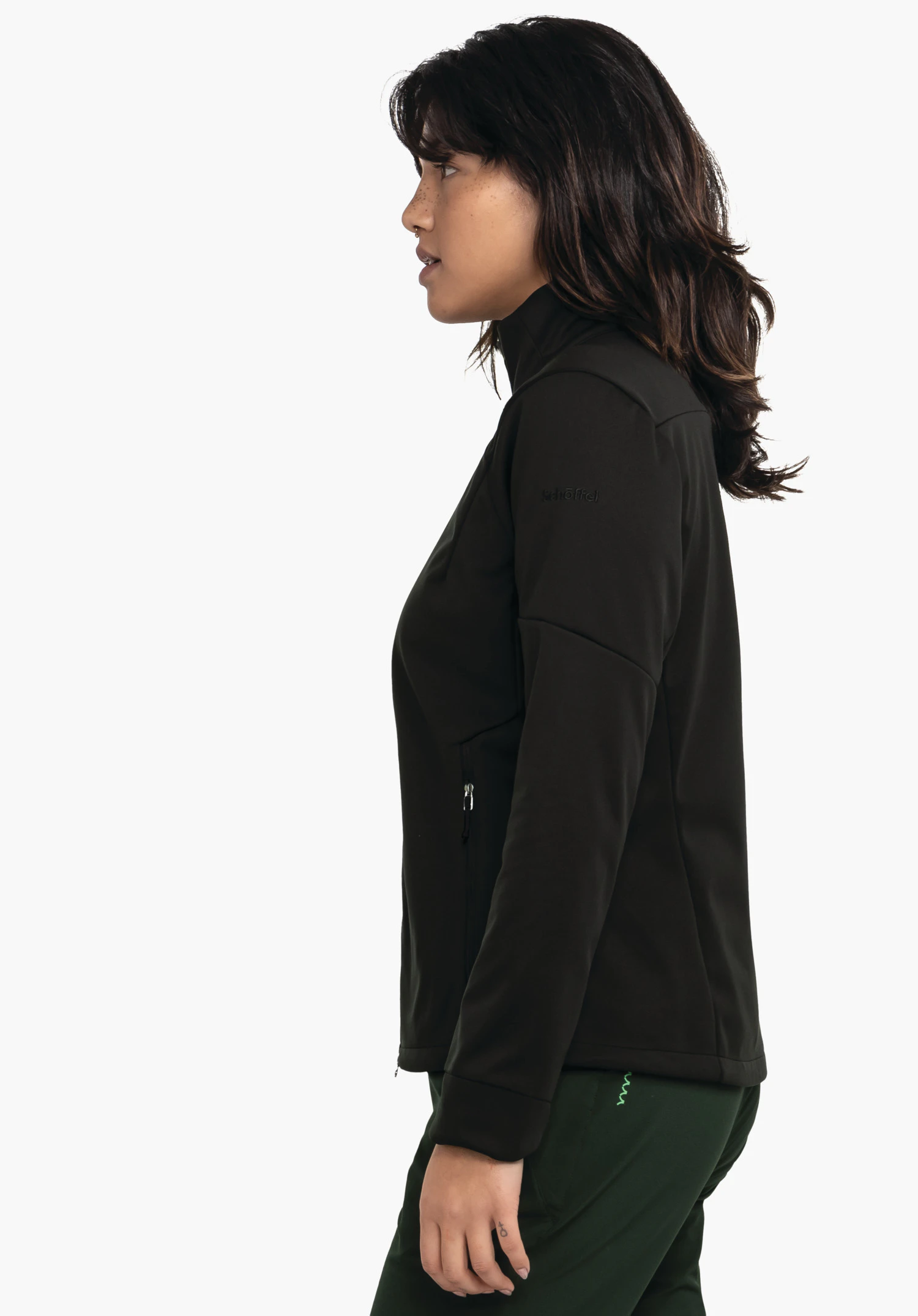 Softshell Jk Style Fracon WMS diepzwarte jas, gedragen door een vrouw in profiel die zijwaarts staat, toont gesloten rits en zak boven een donkergroene broek, voor een lichte witte achtergrond. Tekst: "Schöffel".