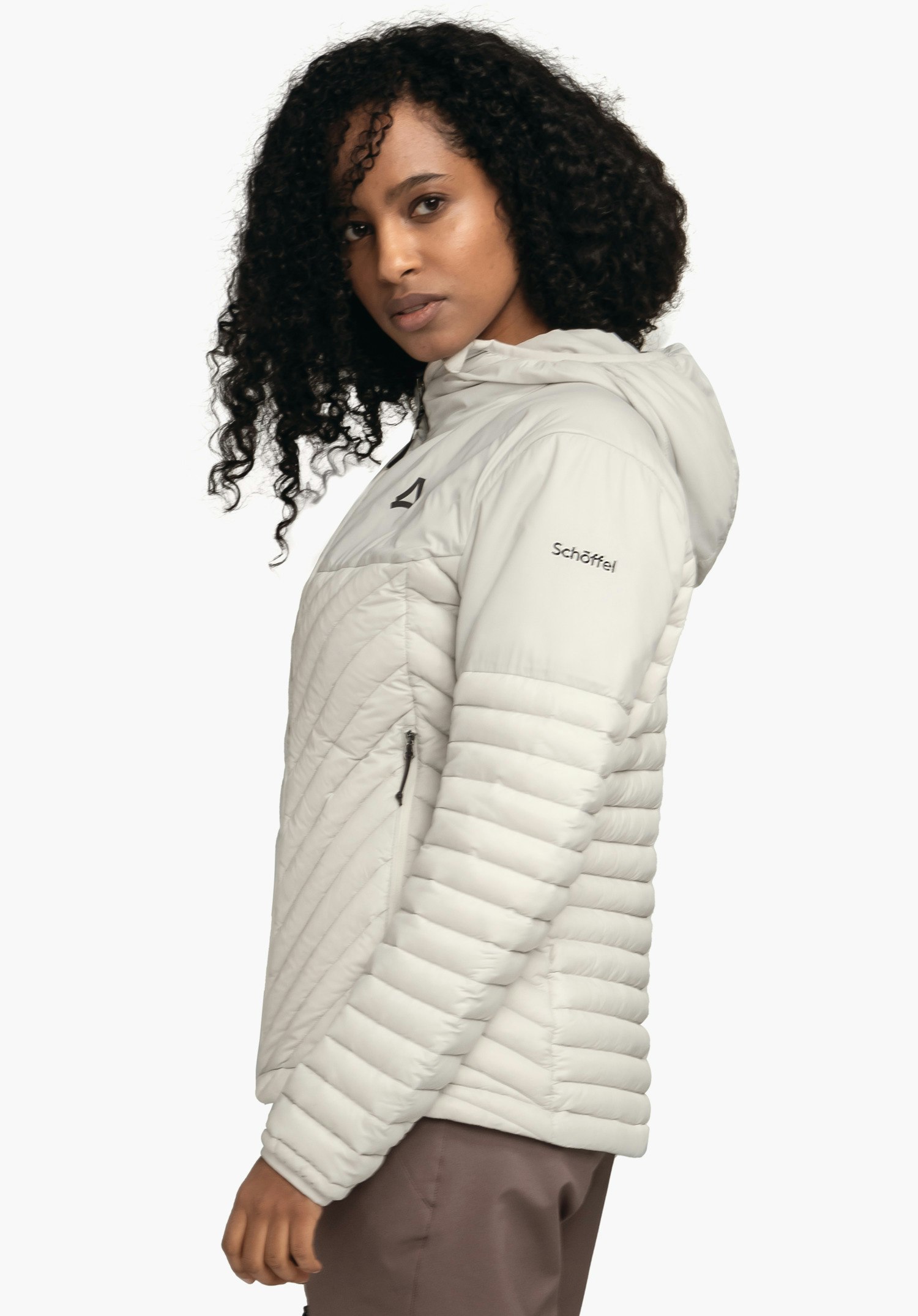 Down Jacket Style Cascata WMS hellgraue Steppjacke, mit Kapuze, trägt eine Frau seitlich, Blick zur Kamera; Schöffel-Logo auf der Brust, Schriftzug "Schöffel" auf dem Ärmel, heller Hintergrund.