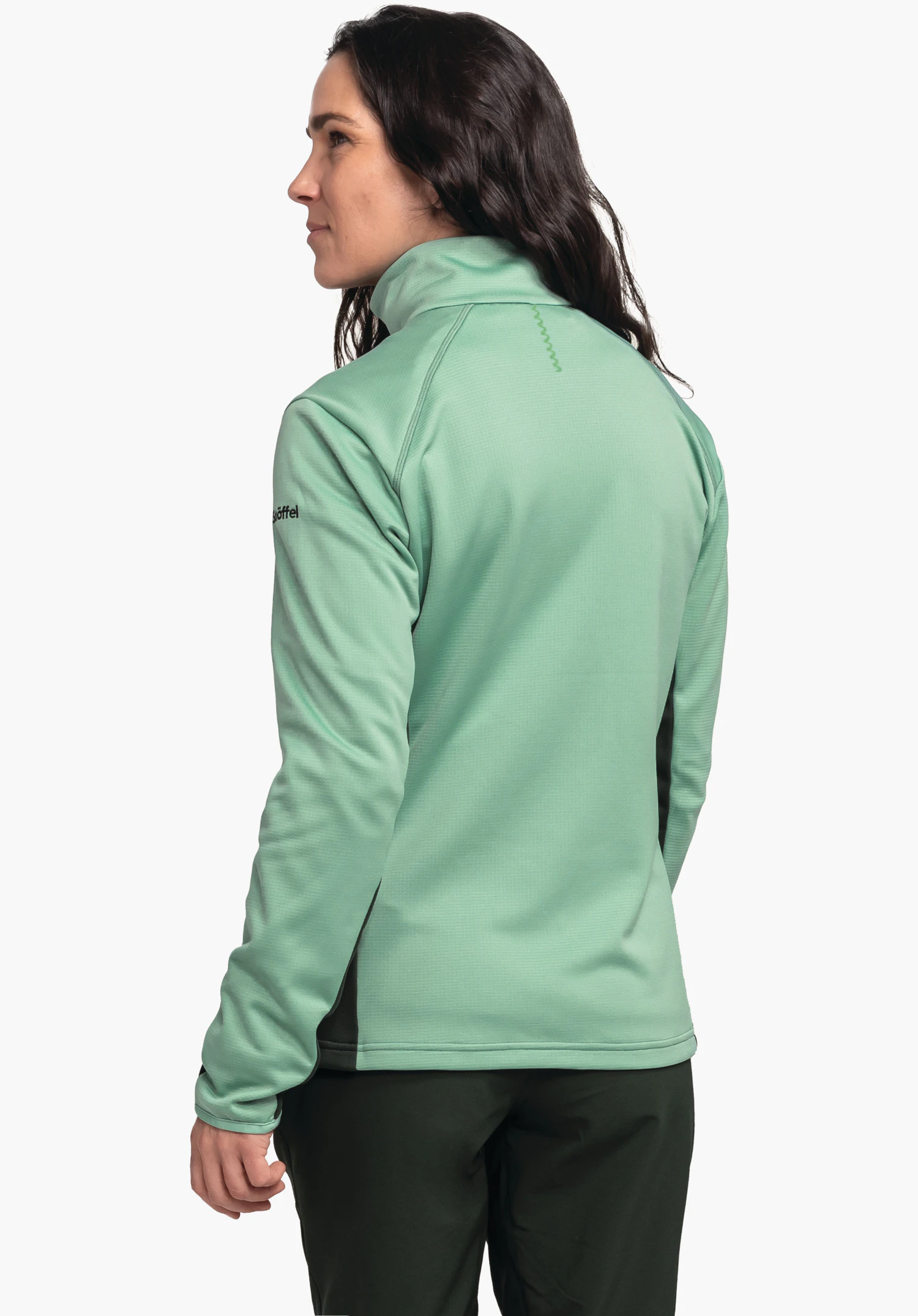 Fleece Jk Style Milagle WMS veste vert clair, portée par une femme debout dos à la caméra et regardant sur le côté; inscription « Schöffel » sur la manche; fond blanc neutre, pantalon vert foncé.