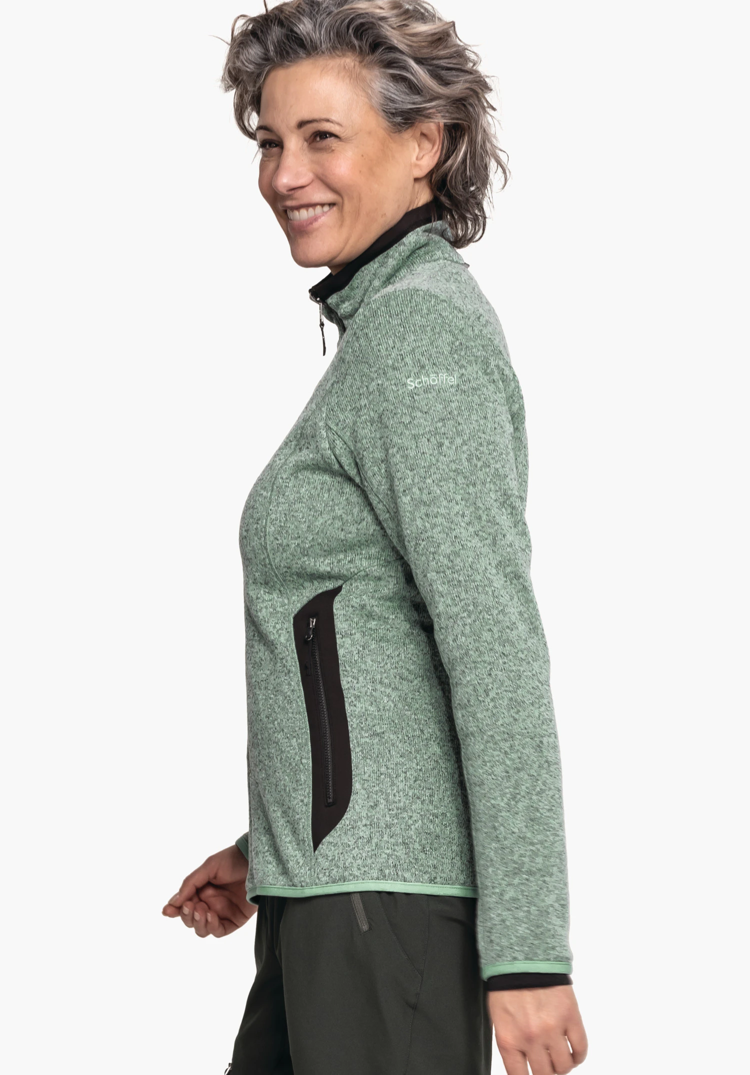 CIRC Fleece Jk Style Yew WMS veste polaire vert clair: personne debout de profil, souriante, portant une veste avec poche zippée noire; pantalon vert foncé; fond blanc. Texte: "Schöffel".