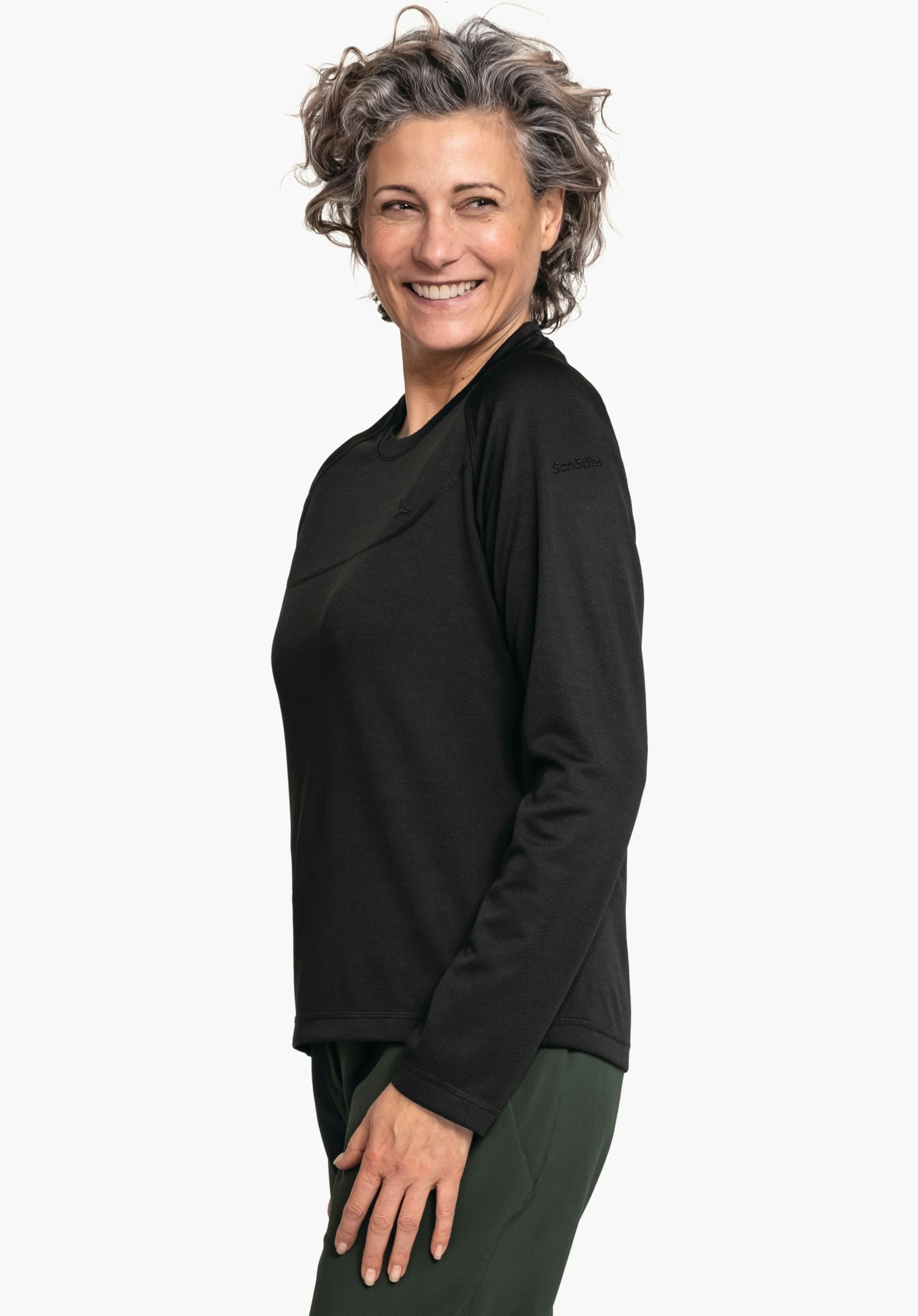 CIRC Longsleeve Smue WMS Eine lächelnde Frau trägt ein langärmliges schwarzes Shirt, steht seitlich vor weißem Hintergrund; Hose dunkelgrün. Text auf Ärmel: "Schöffel".