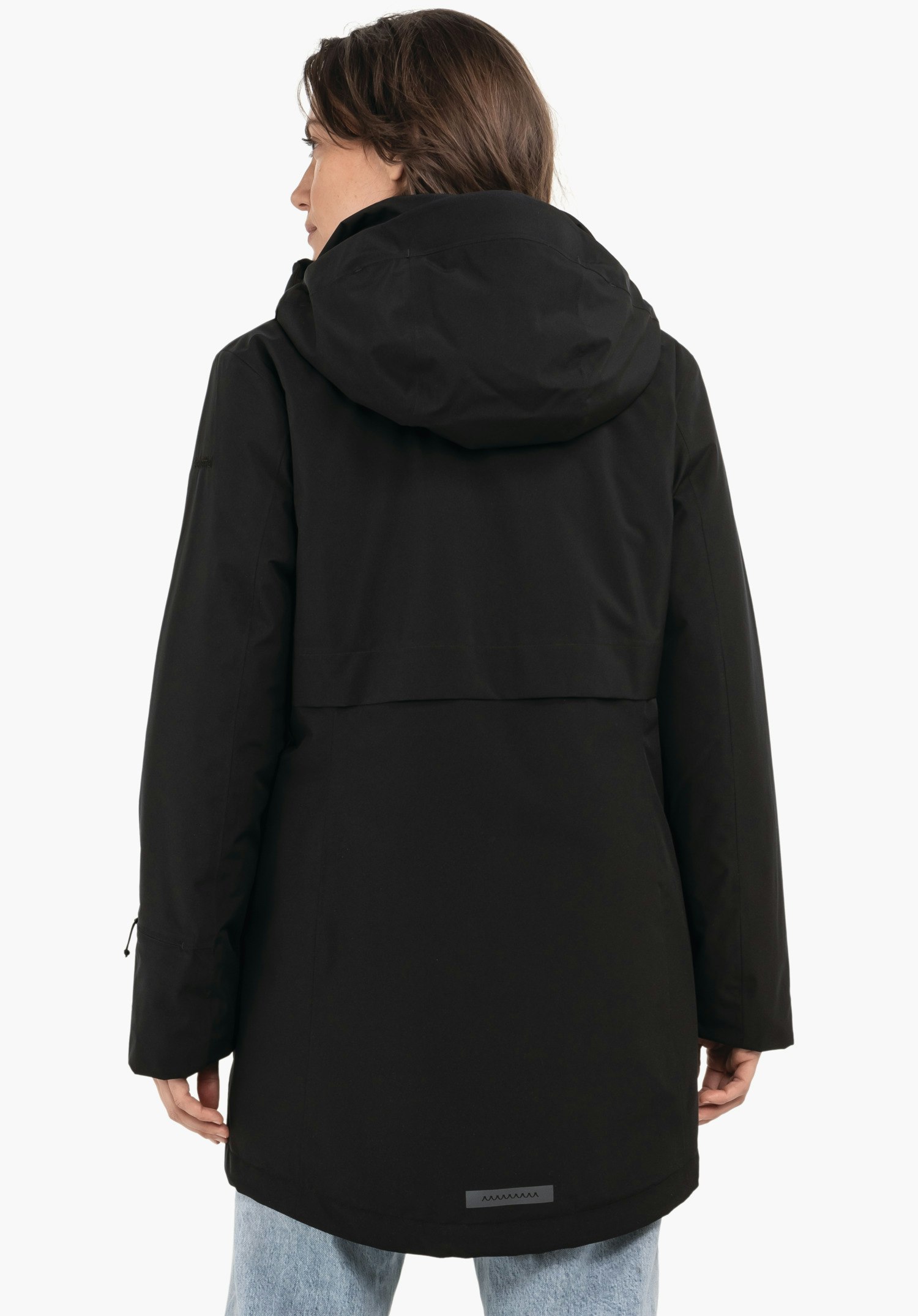 Ins Parka Style Malkay WMS parka noire, porté de dos; personne debout, tête tournée sur le côté; capuche reposant dans le dos, parka couvrant les hanches, jean bleu clair, fond blanc.