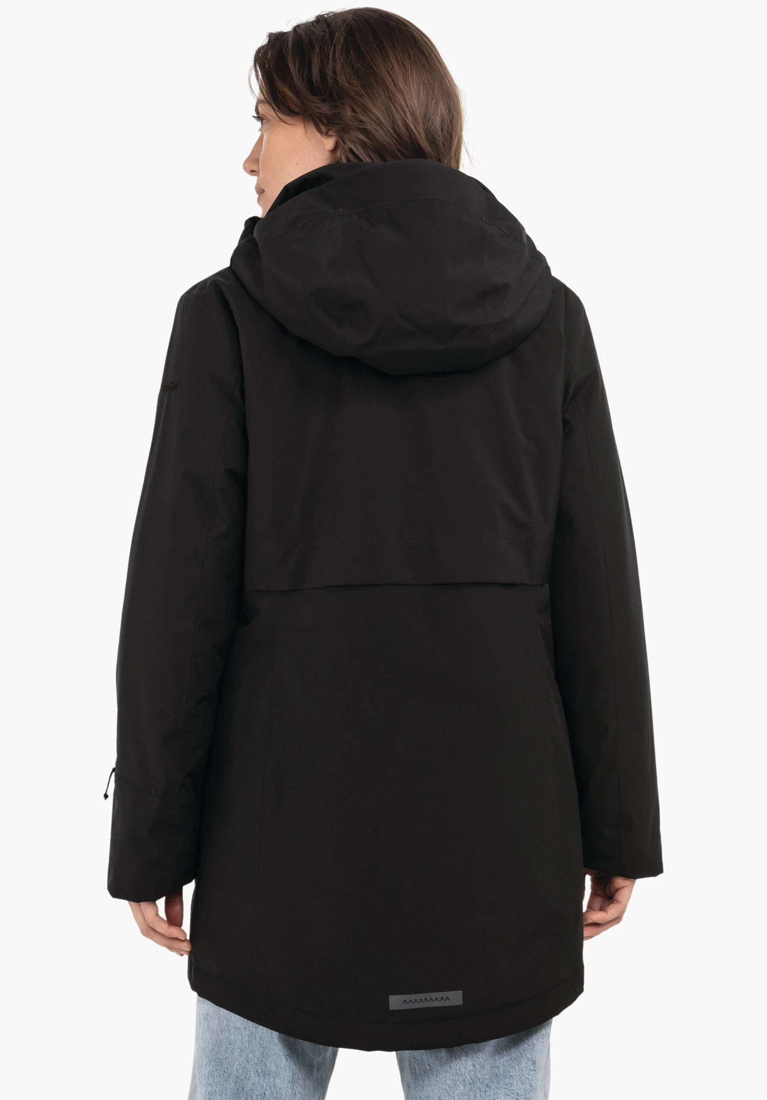 Ins Parka Style Malkay WMS zwarte parka, van achteren gedragen; persoon staat en draait het hoofd opzij, capuchon ligt op de rug, parka reikt over de heupen, lichtblauwe jeans, witte achtergrond.