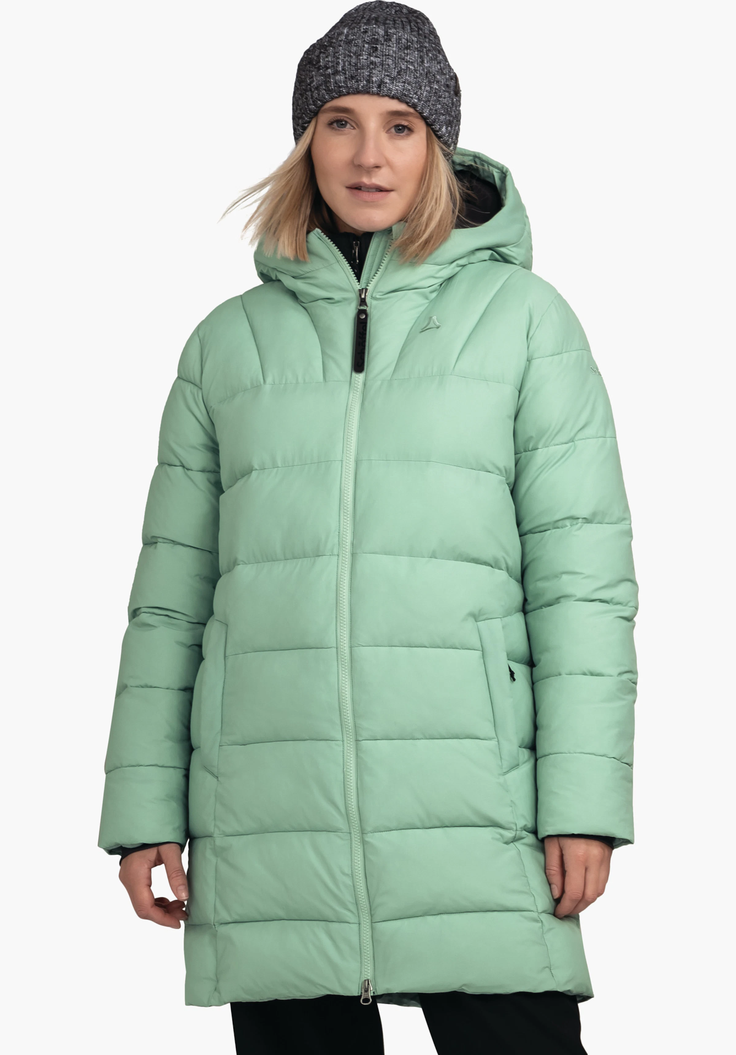 Ins Parka Style Boslix WMS Veste matelassée vert clair portée de face par une femme avec capuche relevée; debout devant un fond blanc; bonnet tricoté gris foncé, bras détendus; logo Schöffel sur la poitrine, marquage «Schöffel» sur le zip.