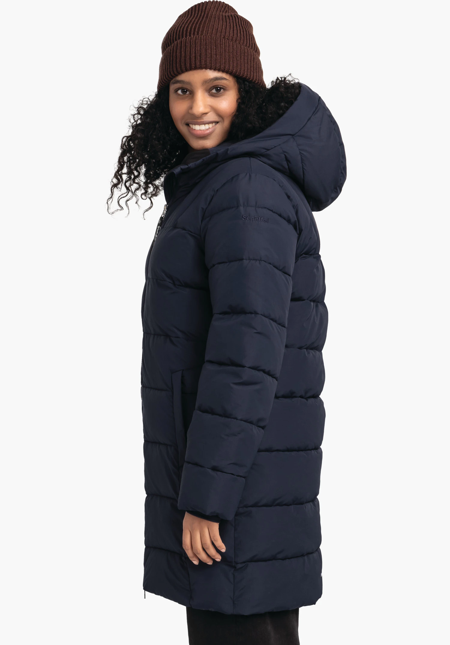 Ins Parka Style Boslix WMS donkerblauwe gewatteerde parka met capuchon, zijwaarts gedragen door een glimlachende vrouw; zij draagt een donkerbruine muts, witte achtergrond. Op de mouw: "Schöffel".