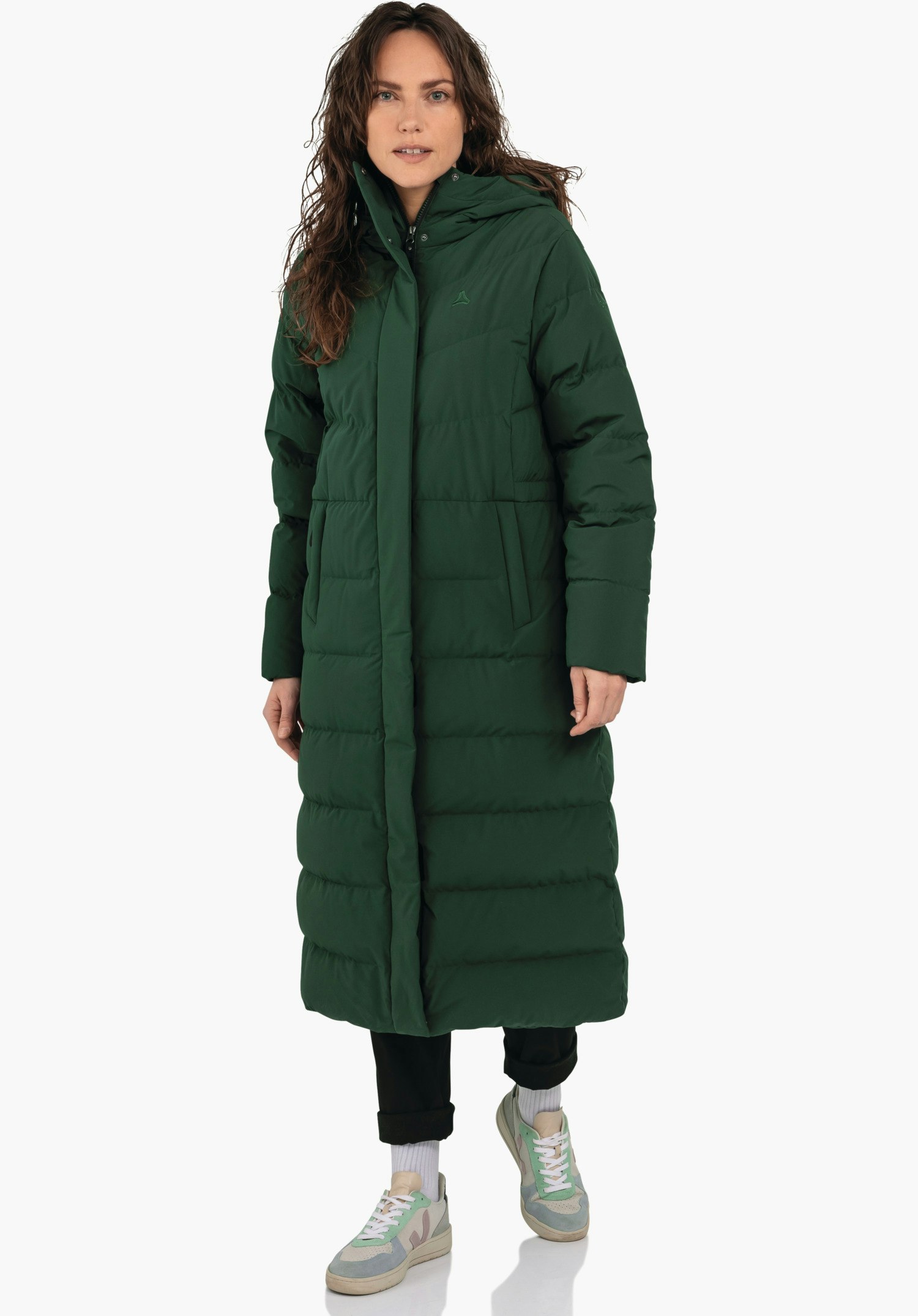 Down Coat Style Vindave WMS dark green long quilted coat worn by a standing woman; hood, side pockets, Schöffel logo on the chest; white background, black trousers, light green and light blue trainers.
